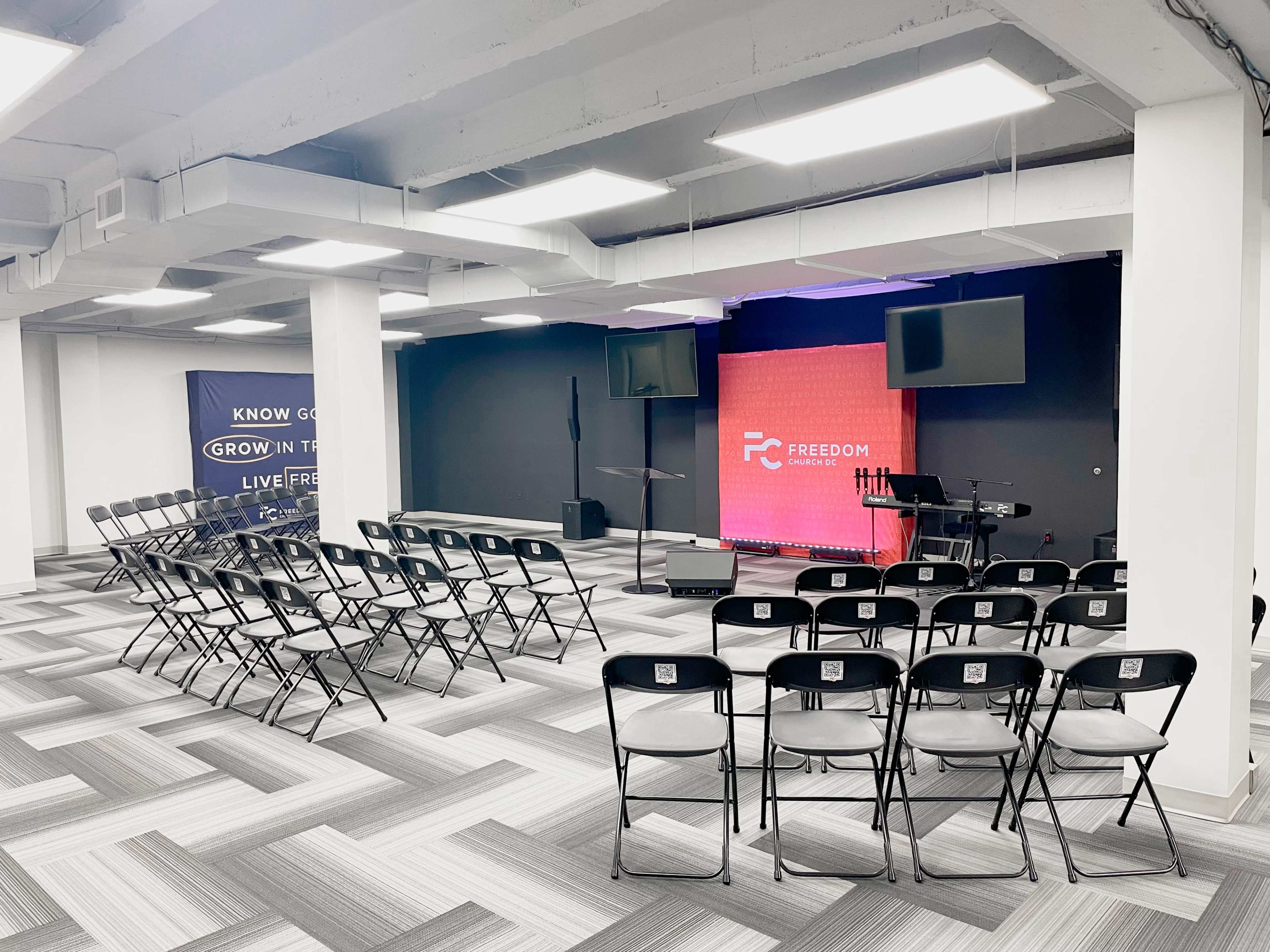 A spacious room set up for an event, with rows of black chairs facing a stage that features a backdrop and musical equipment.