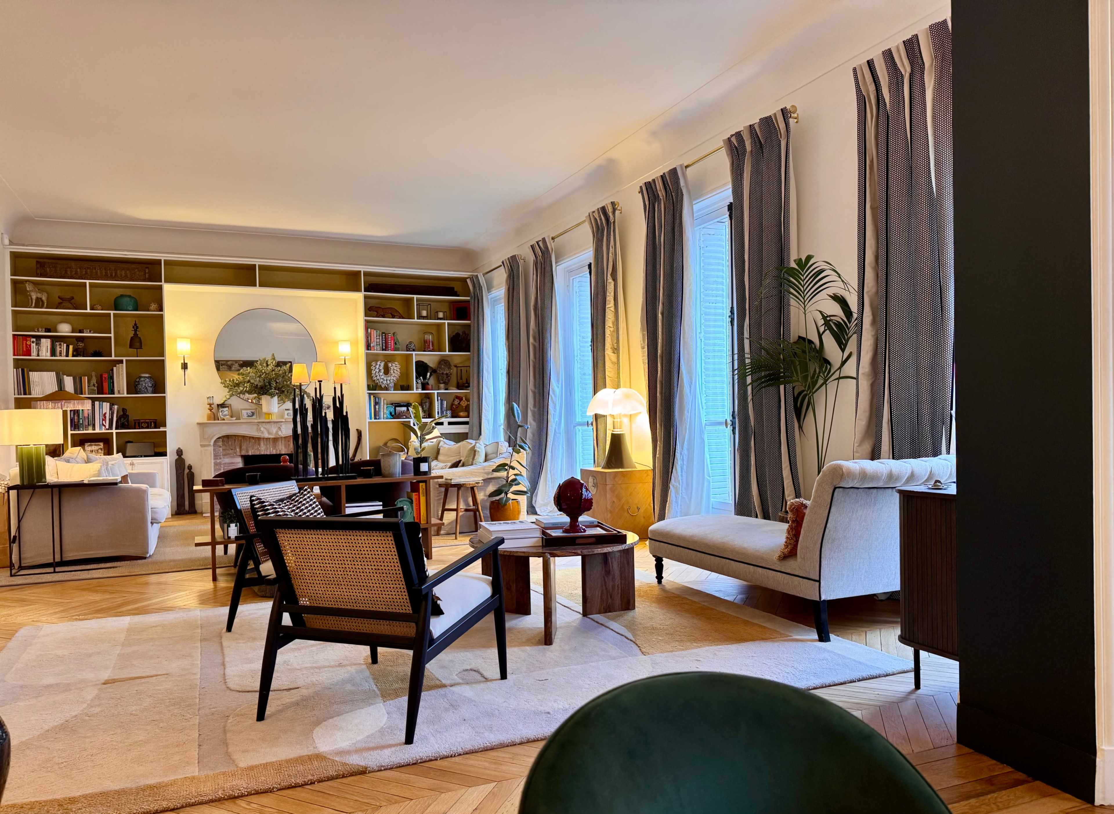 A modern living room features a mix of seating arrangements, bookshelves, and large windows with natural light.