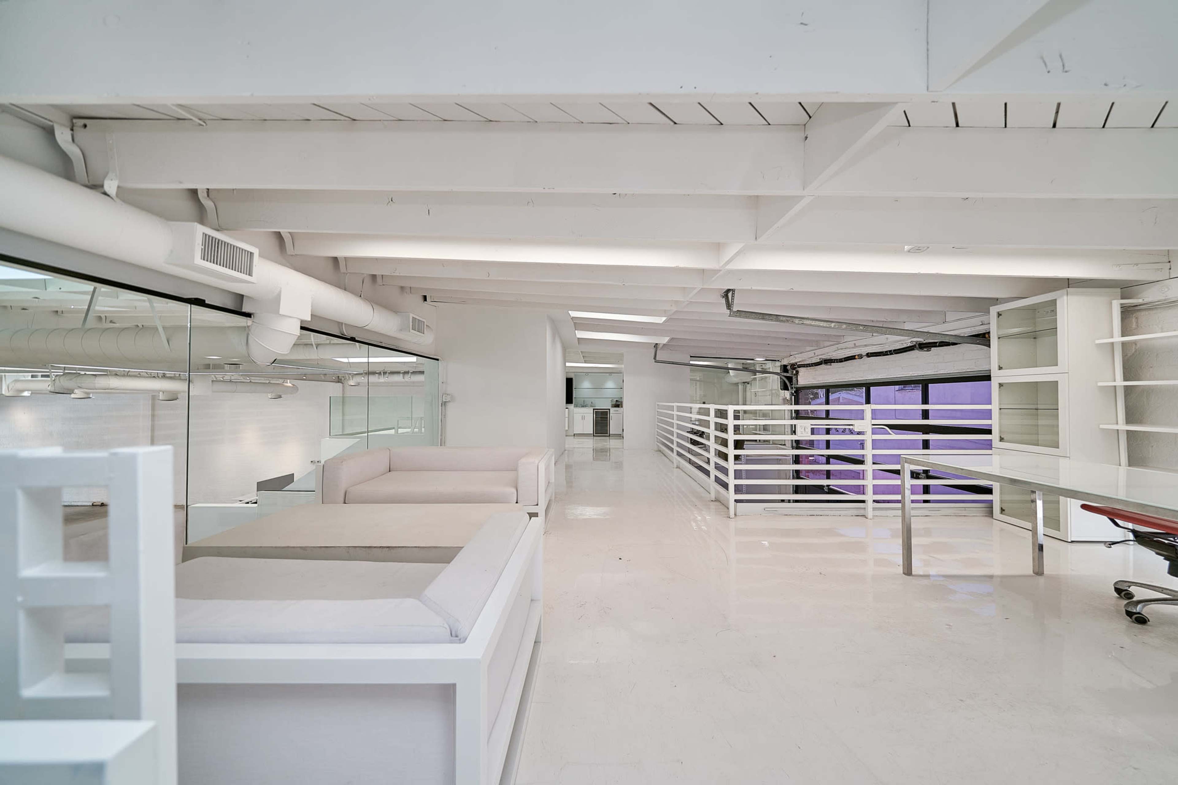 A spacious, modern interior with white walls, exposed beams, and minimal furniture, featuring a seating area and a work desk.
