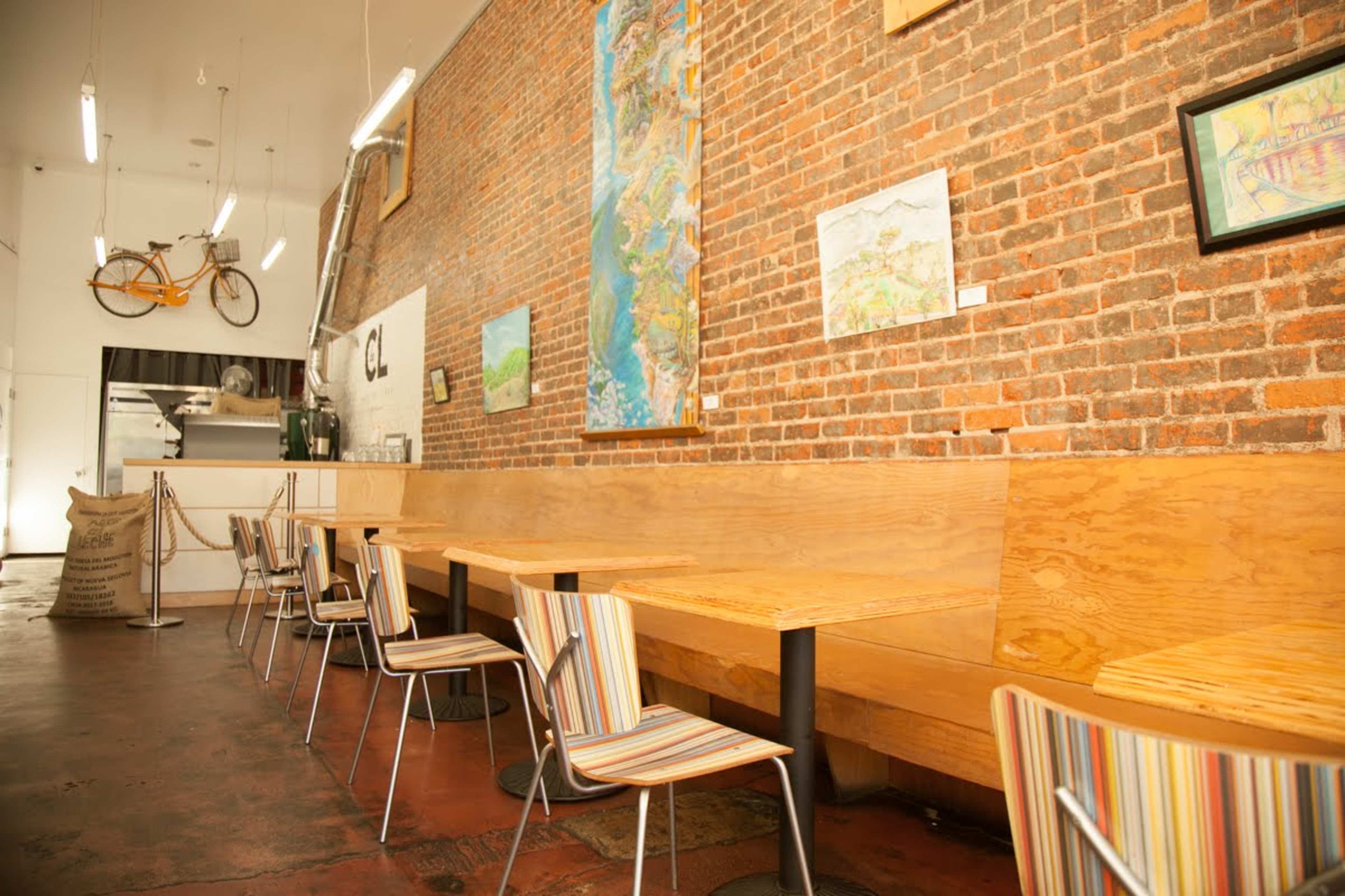 A rustic café interior featuring wooden benches, striped chairs, and a brick wall adorned with artwork and a bicycle.