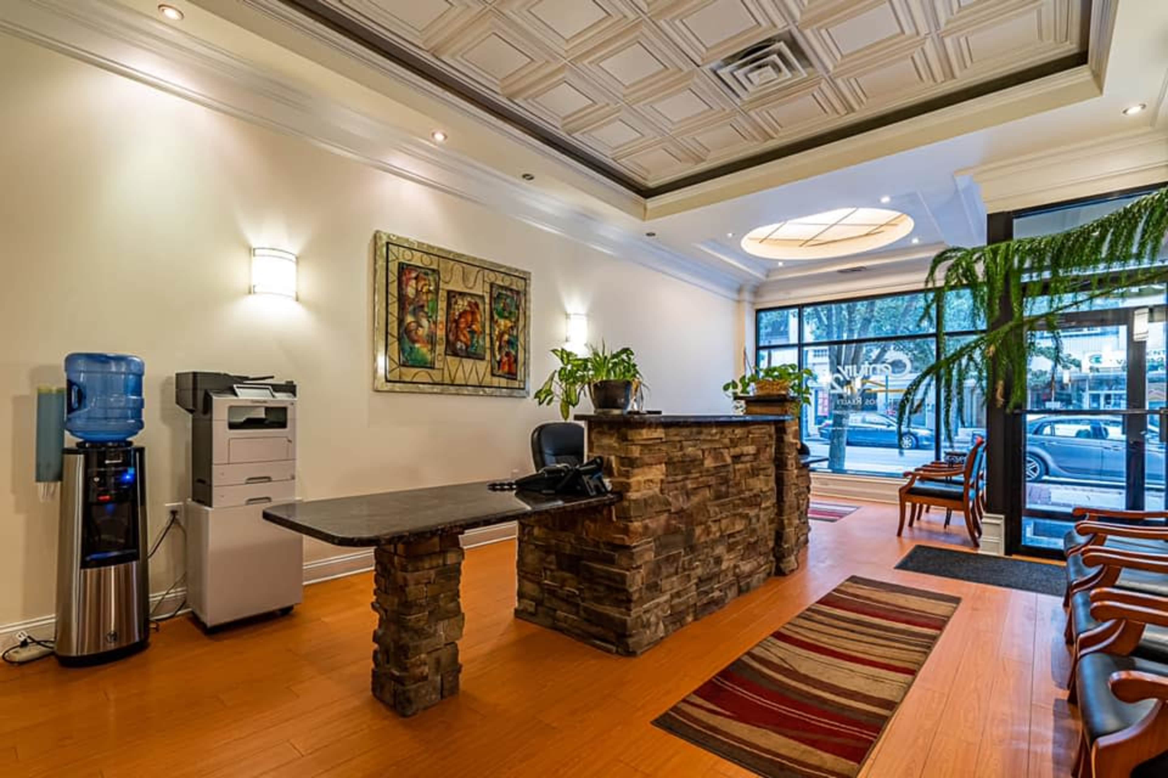 The image shows a modern reception area with a stone front desk, a printer, a water cooler, and potted plants, designed with wooden flooring and large windows.