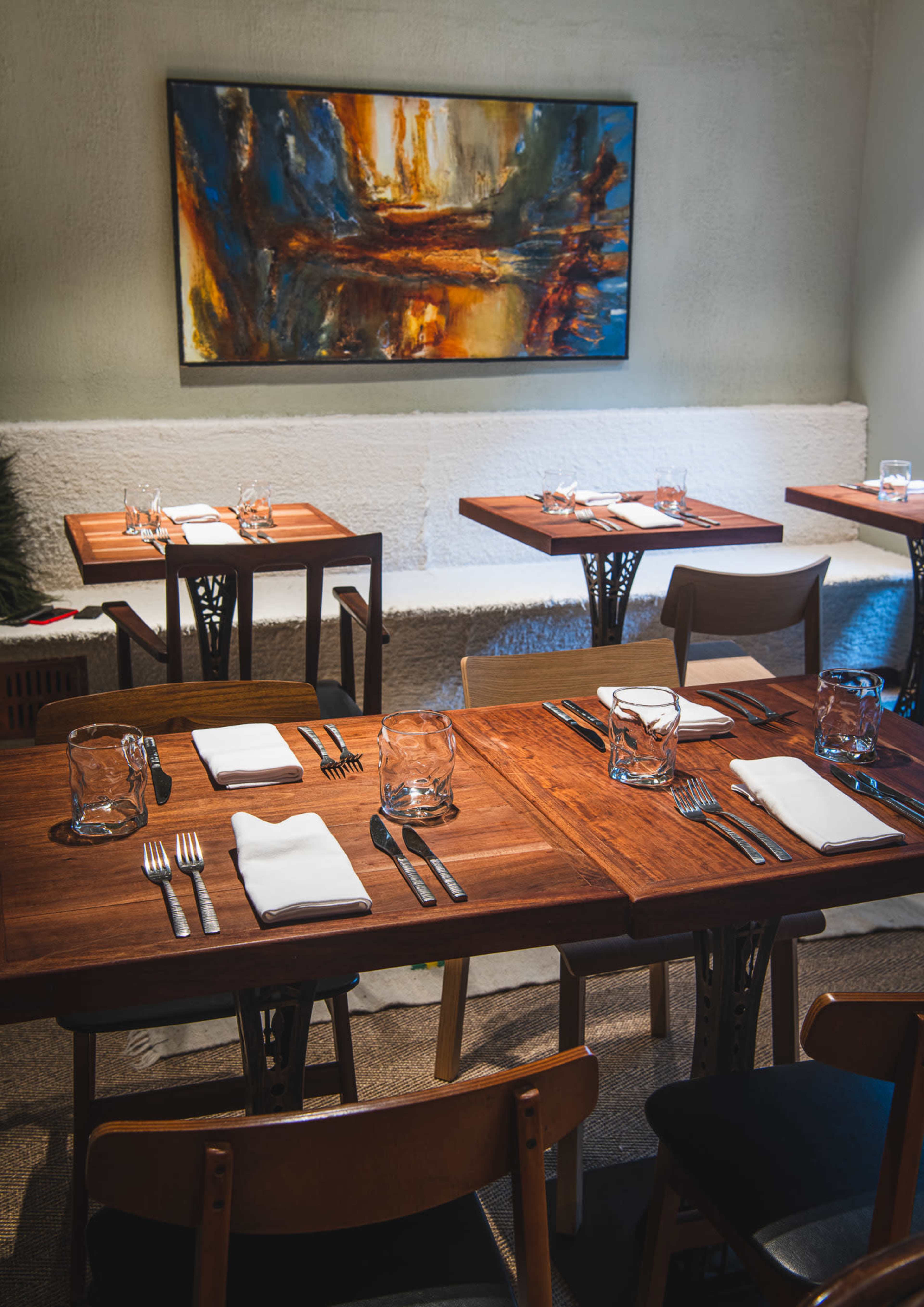 The image shows a dining area with wooden tables set for meals, featuring cutlery and glassware, against a backdrop of an abstract painting.