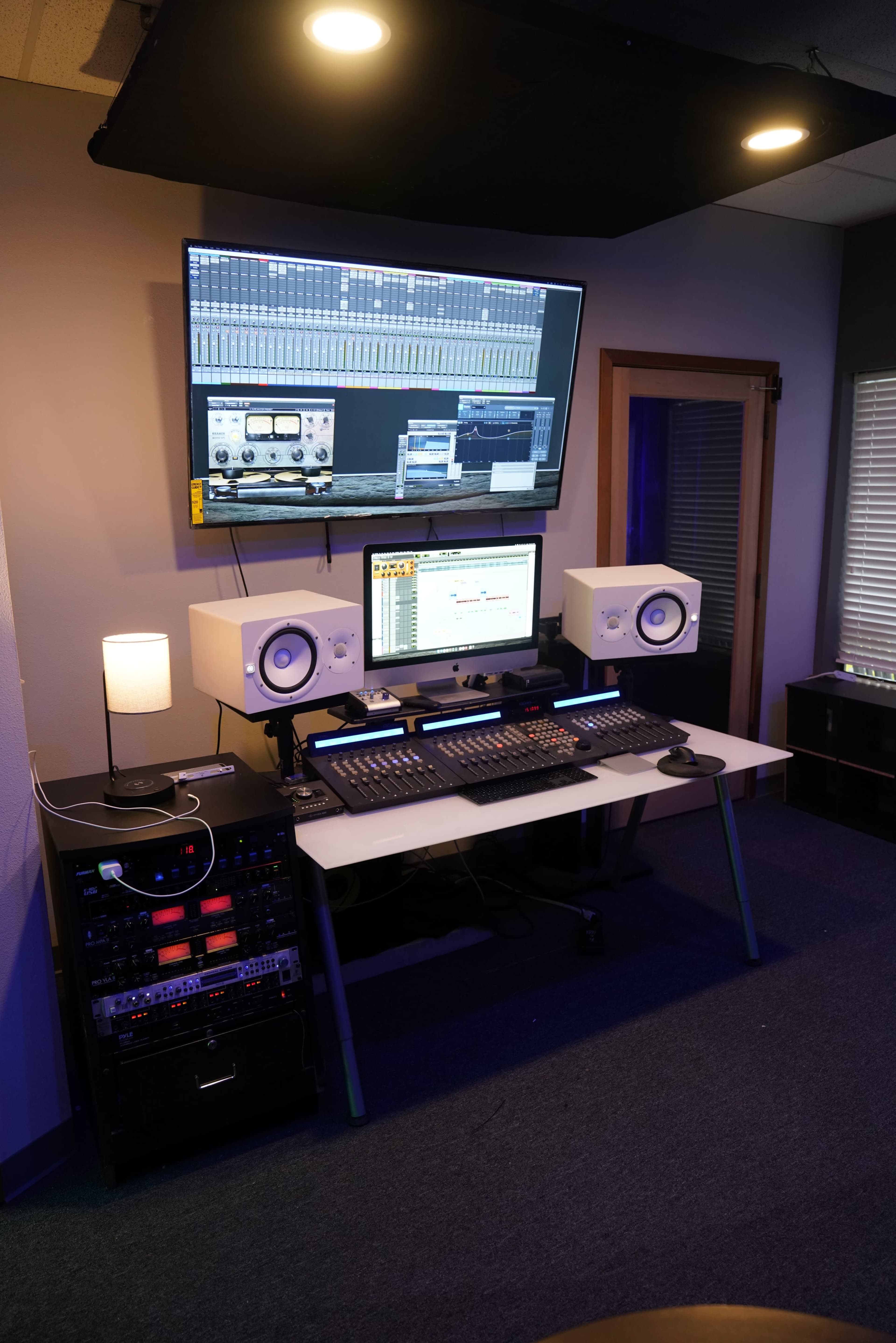 A modern audio recording studio setup with a computer, audio interface, monitors, and a large wall-mounted screen displaying audio software.