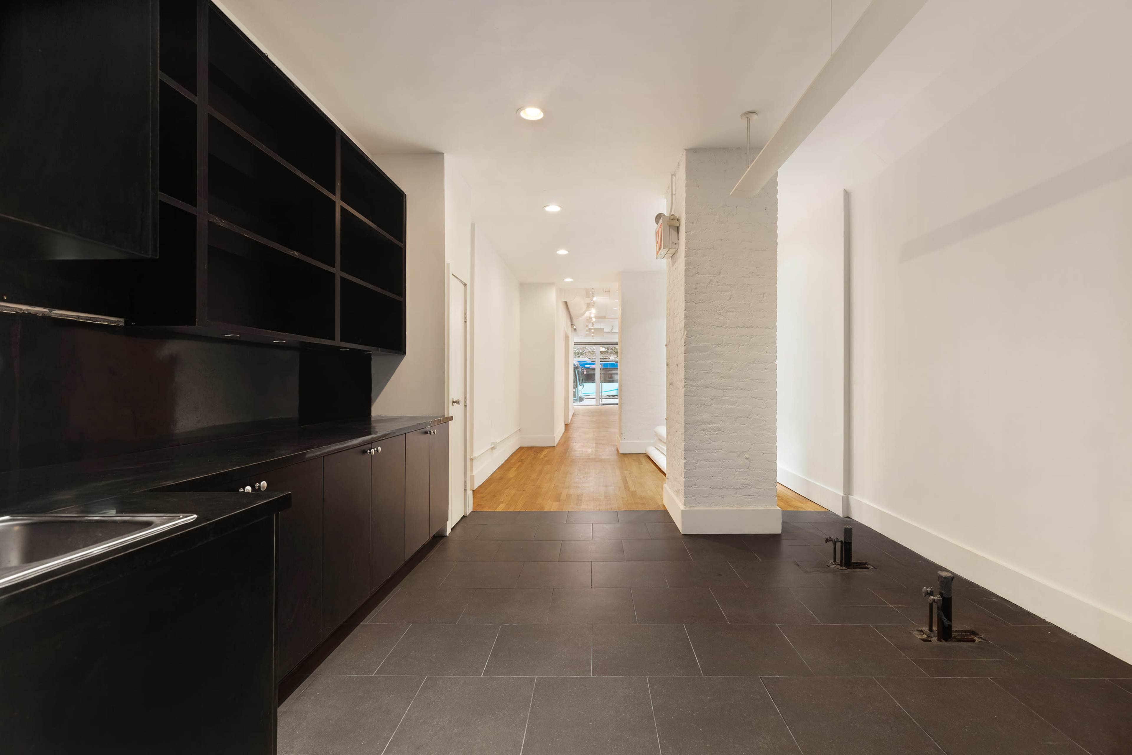 The image shows a narrow hallway leading into a brightly lit space, featuring dark cabinets on one side and light-colored walls.