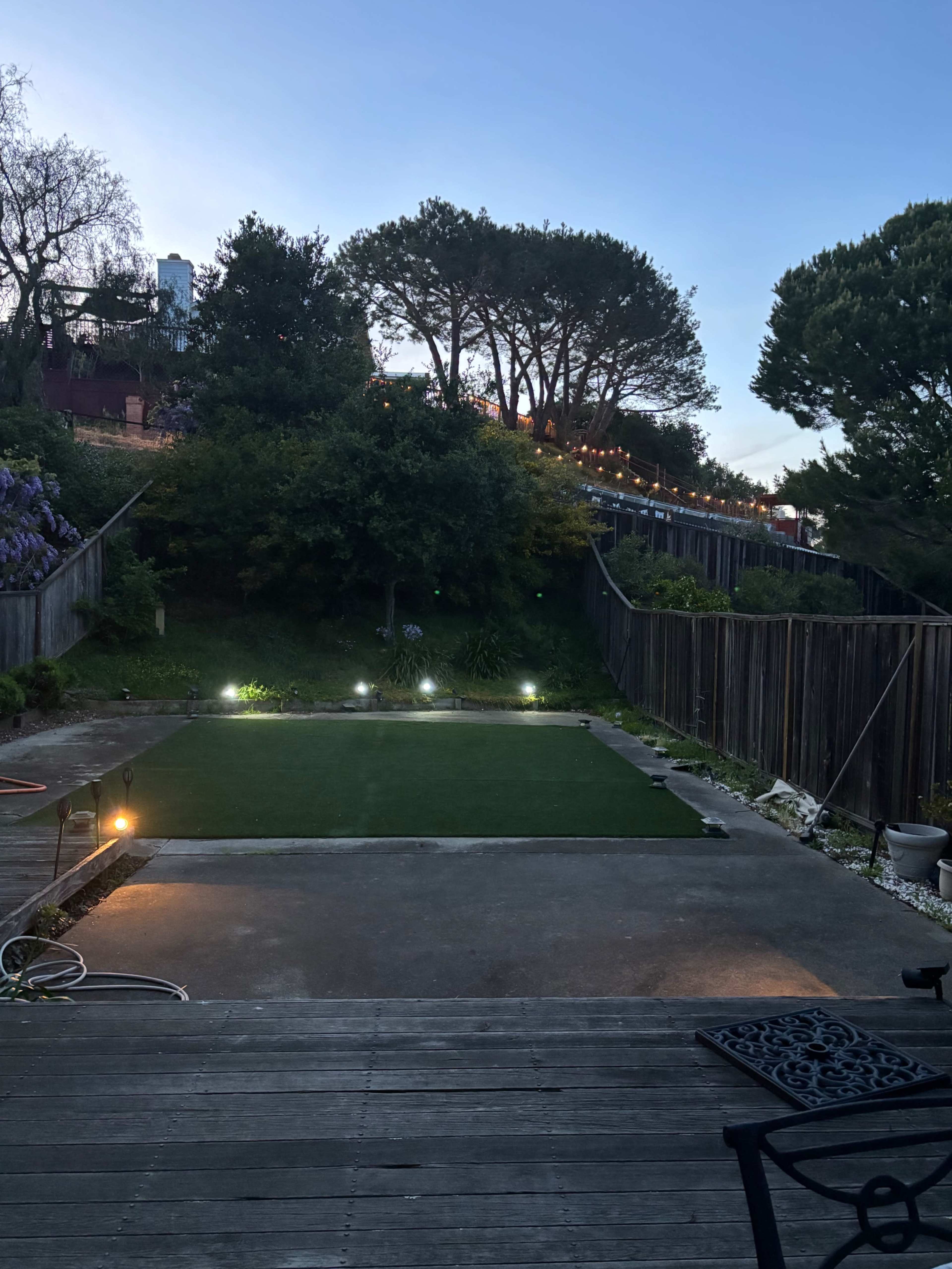 The image shows a backyard with a green lawn area surrounded by wooden fences and trees, illuminated by soft lights in the evening.