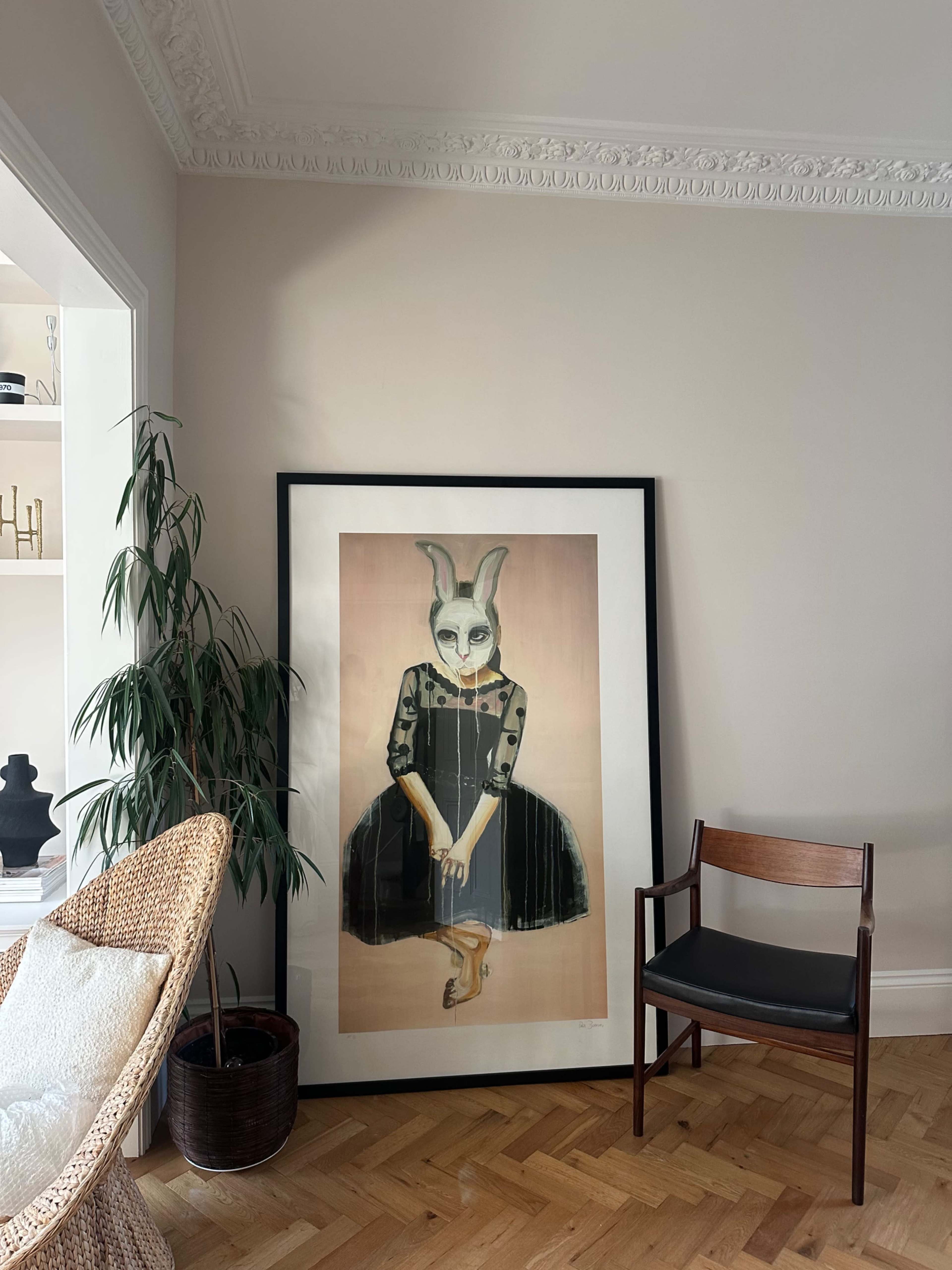 The image features a large framed artwork of a figure with a rabbit mask wearing a black dress, positioned beside a potted plant and a chair in a well-lit room.