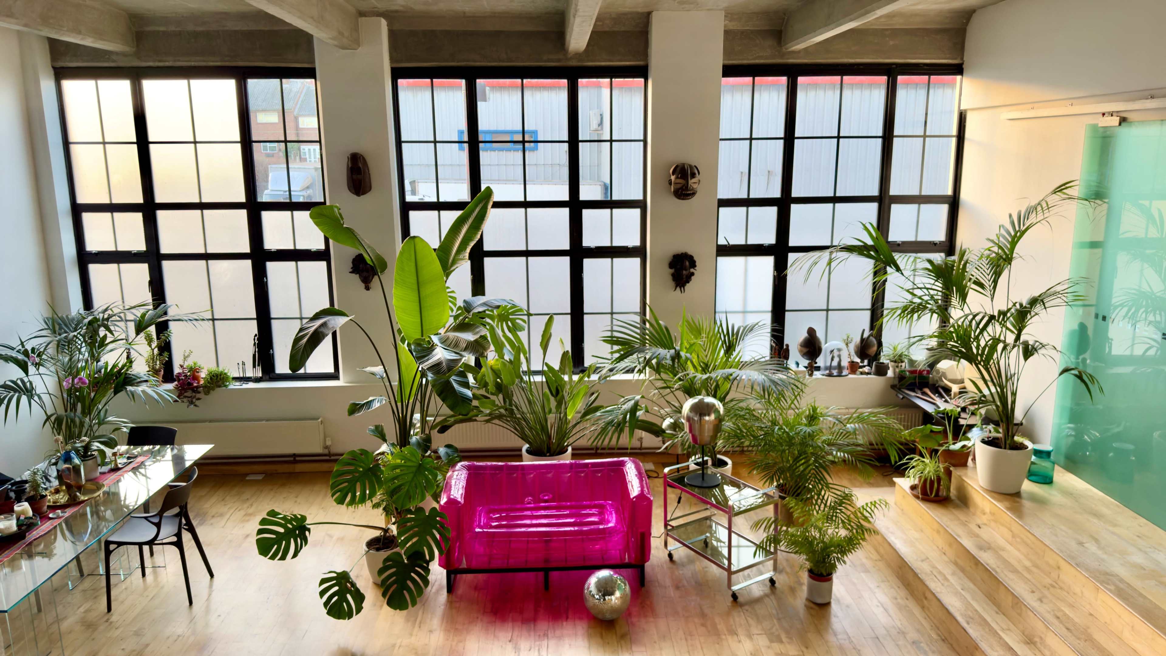 The image shows a spacious interior with large windows, featuring a pink couch surrounded by various indoor plants and modern decor.