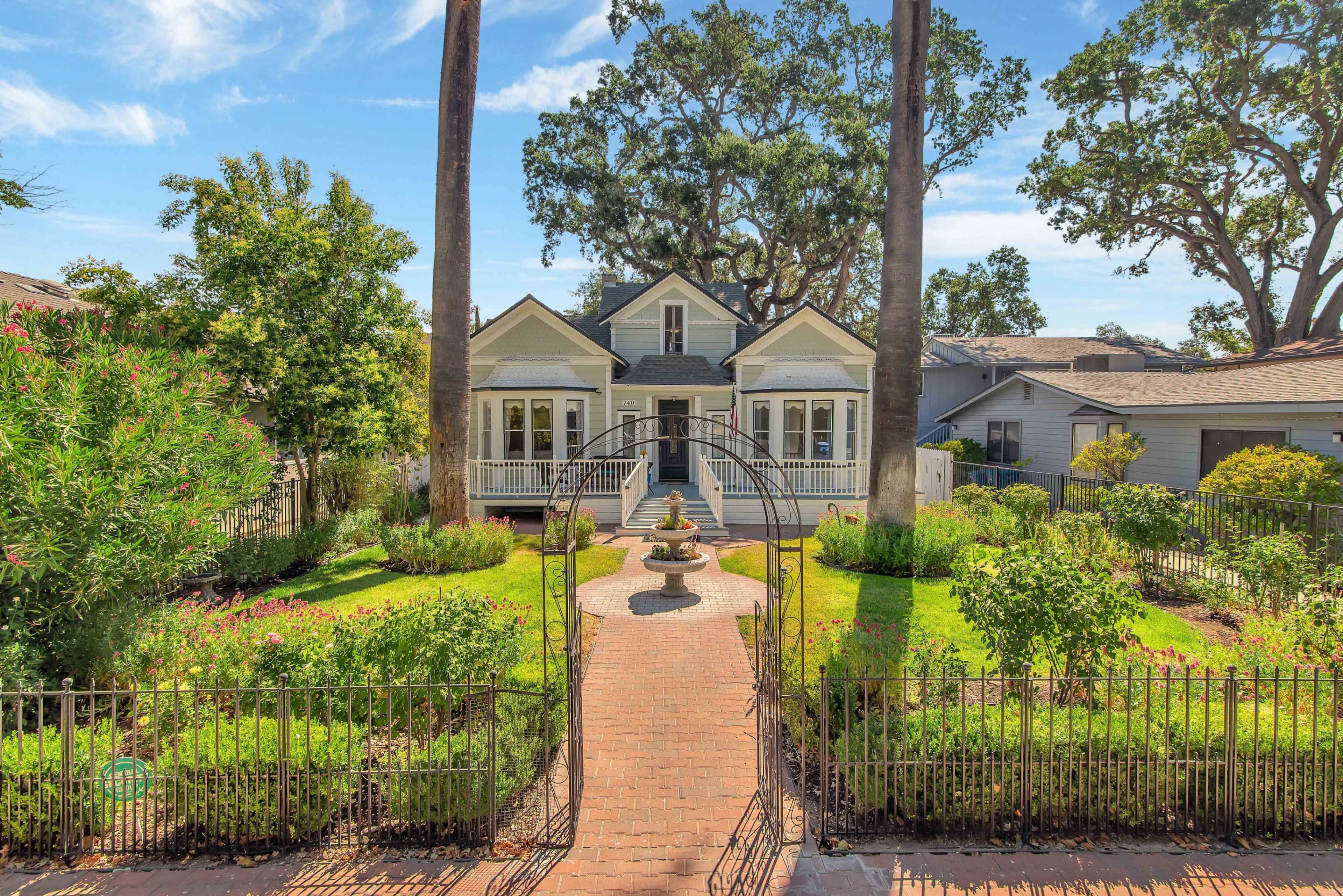 A two-story house with a front porch is surrounded by a well-maintained garden featuring a central fountain and tall palm trees.