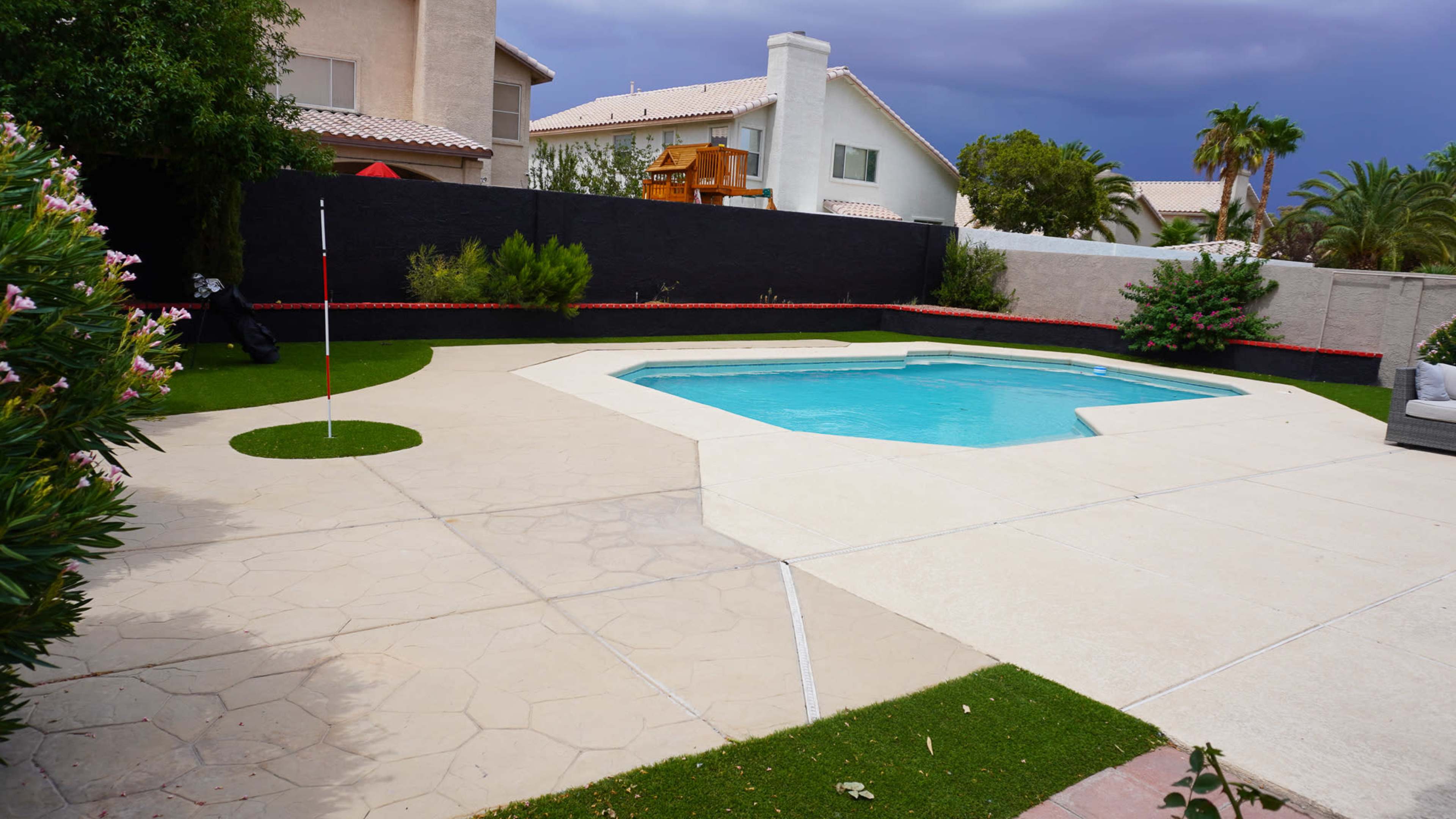 A backyard with a swimming pool surrounded by concrete and a grassy area, featuring a palm tree and a playset in the background.