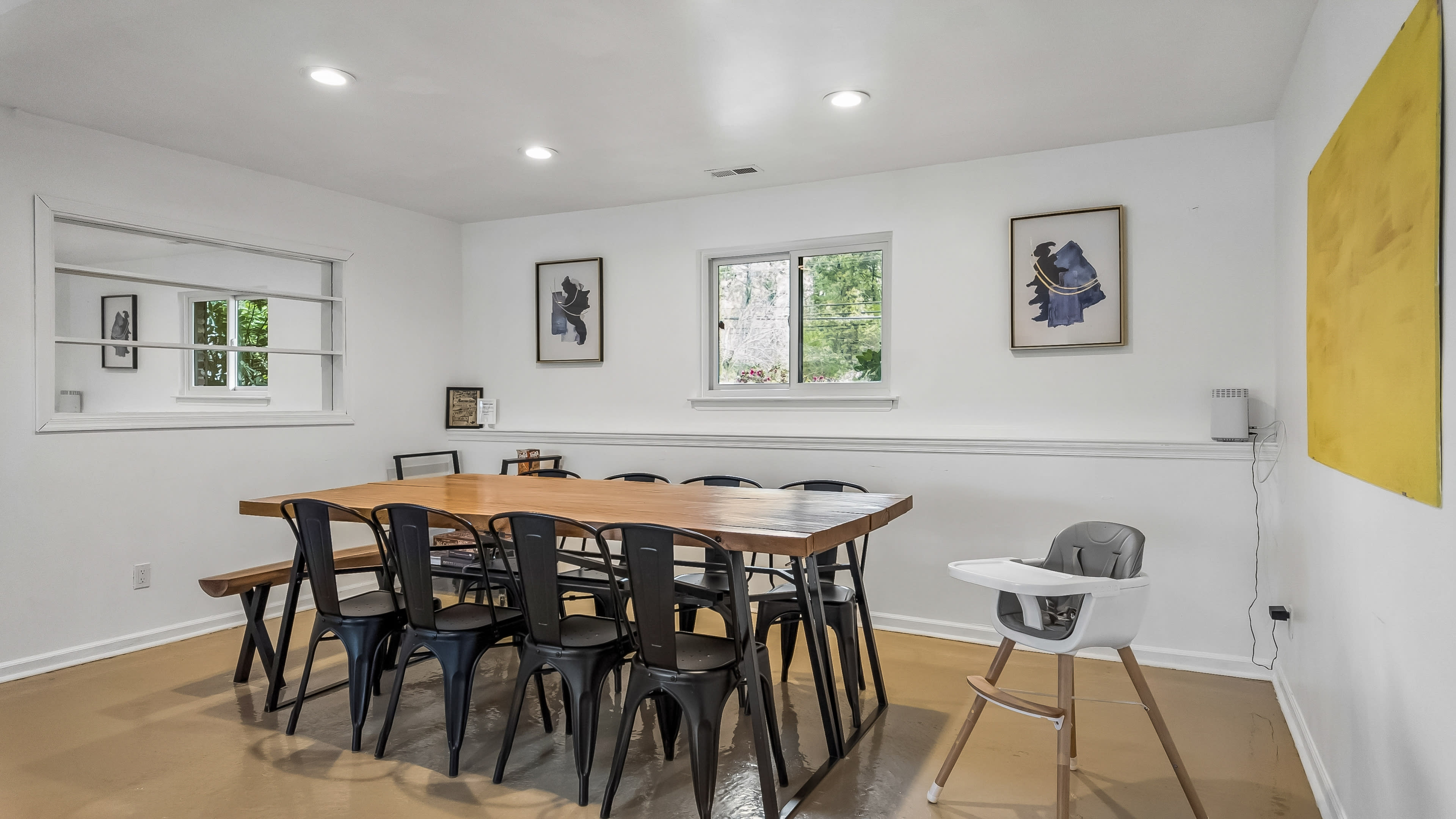 The image shows a modern dining area with a large wooden table surrounded by black chairs, white walls, and framed artwork.