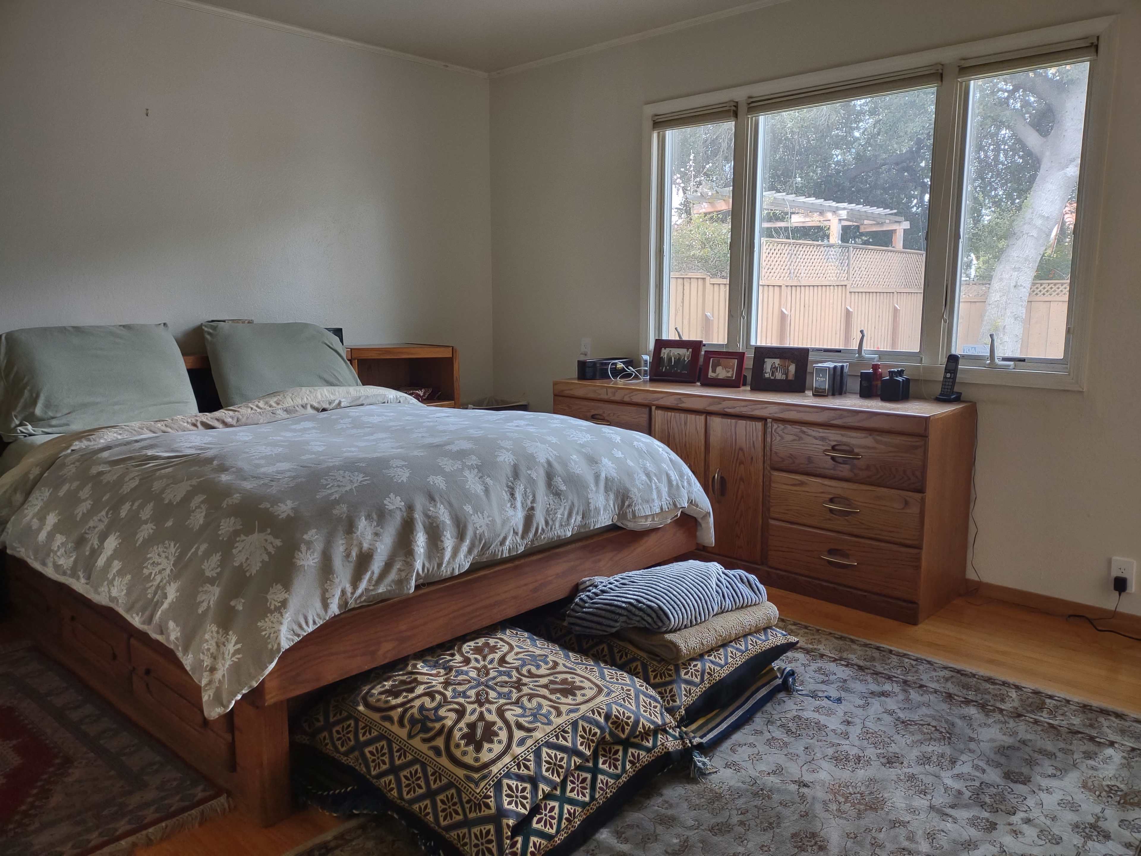 A spacious bedroom features a wooden bed with gray bedding, a dresser against the wall, and large windows overlooking a fenced yard.