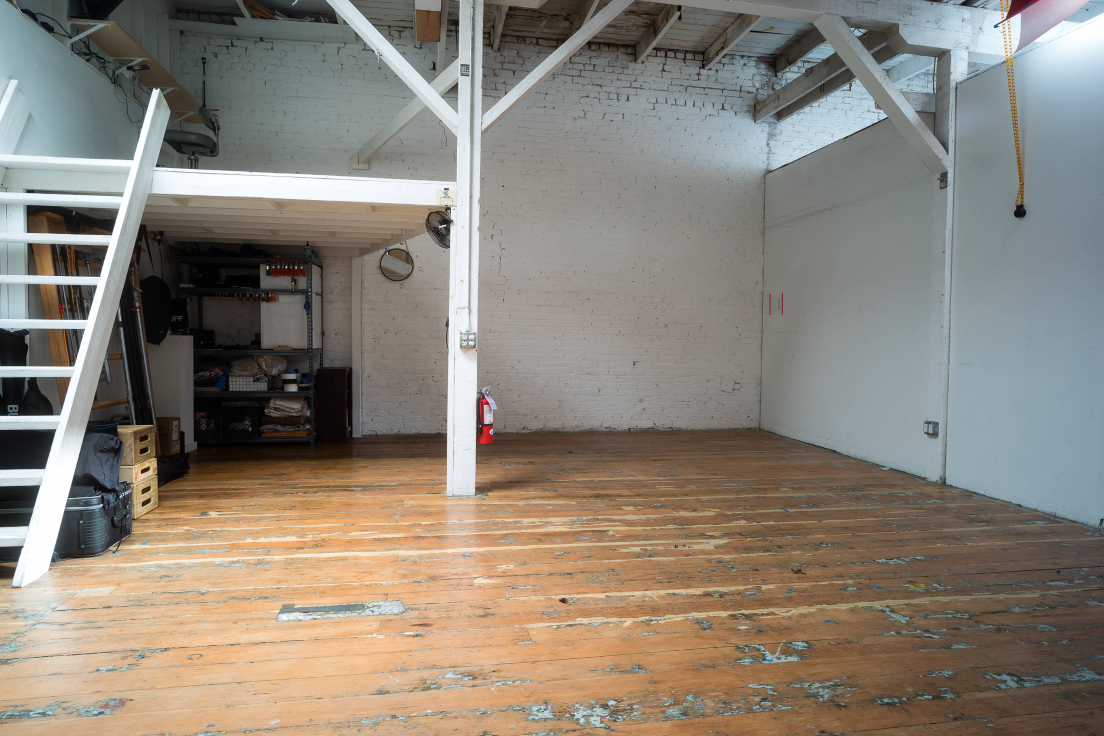 A spacious, empty room with wooden floors, exposed ceiling beams, and a wooden staircase leading to a loft area.