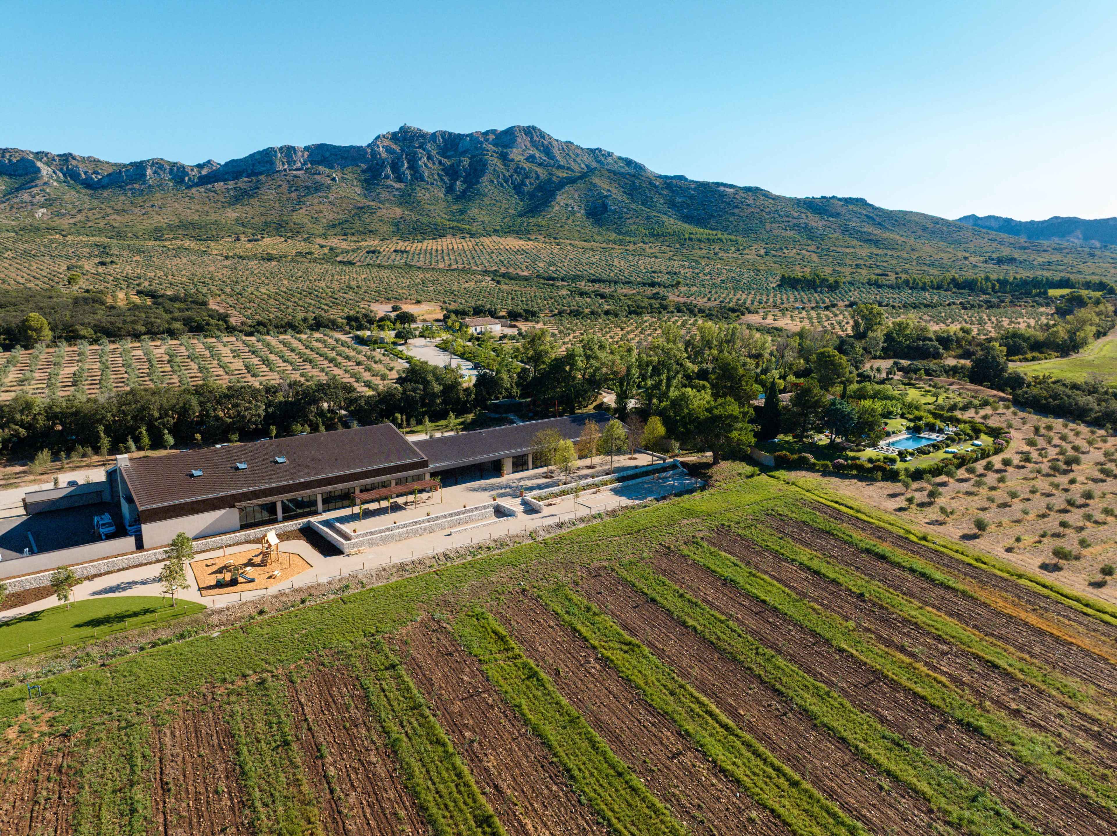 The image shows a large, modern building surrounded by fields and hills, with cultivated rows of crops in the foreground and a mountainous landscape in the background.
