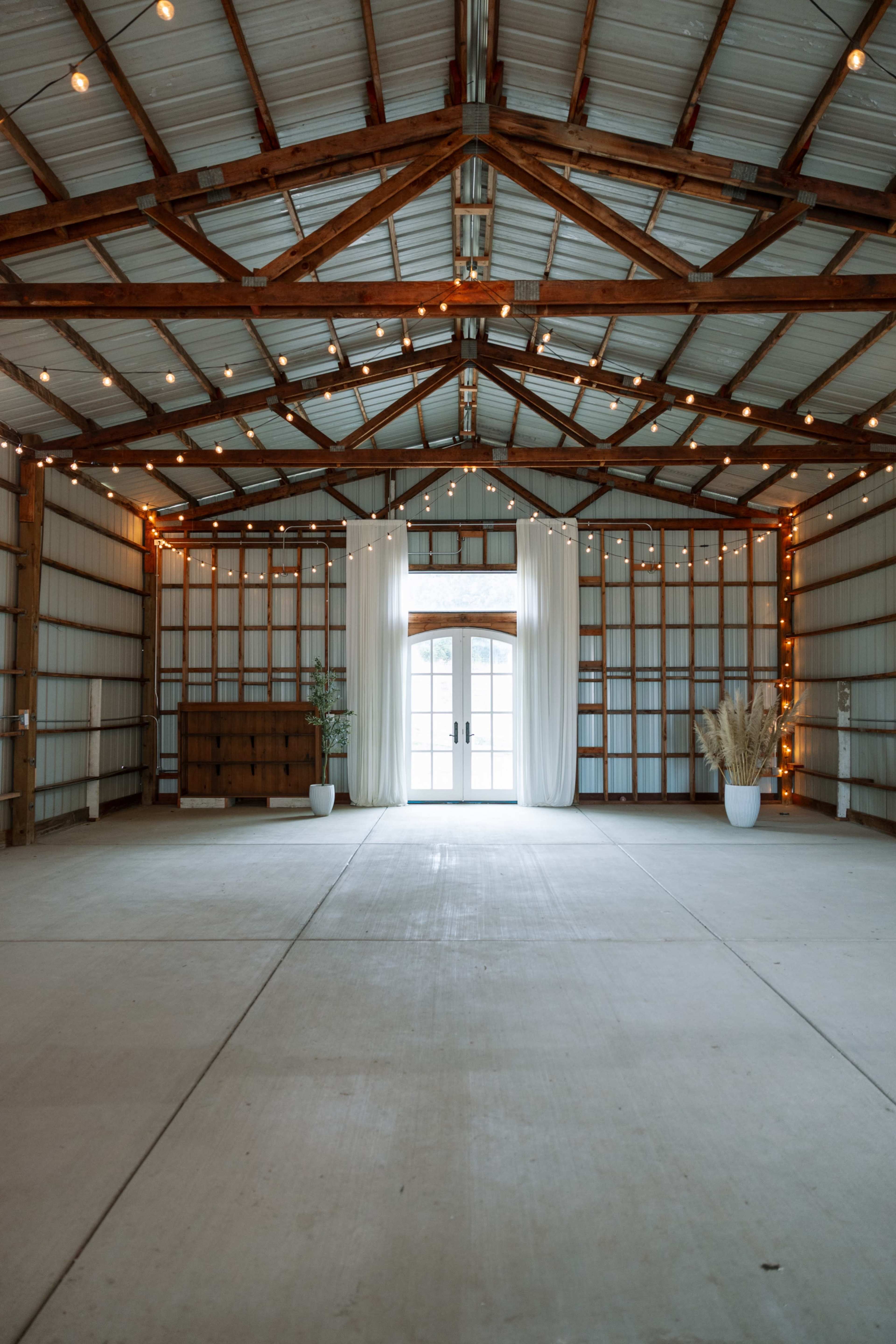 The interior of a barn features wooden beams and string lights, with a couple of potted plants near large double doors.