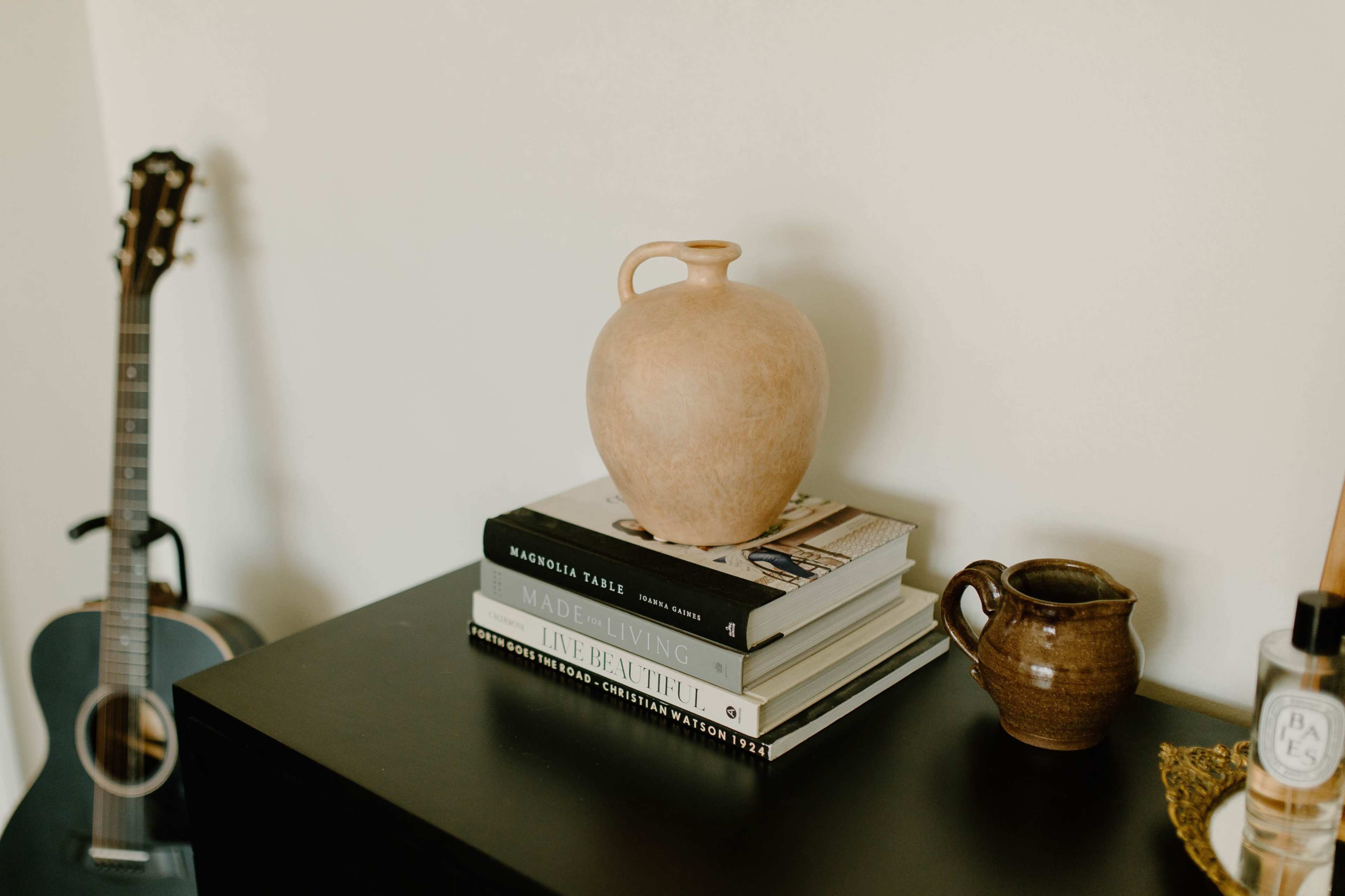 A terracotta vase is placed on top of a stack of books beside a guitar and a small ceramic pitcher.