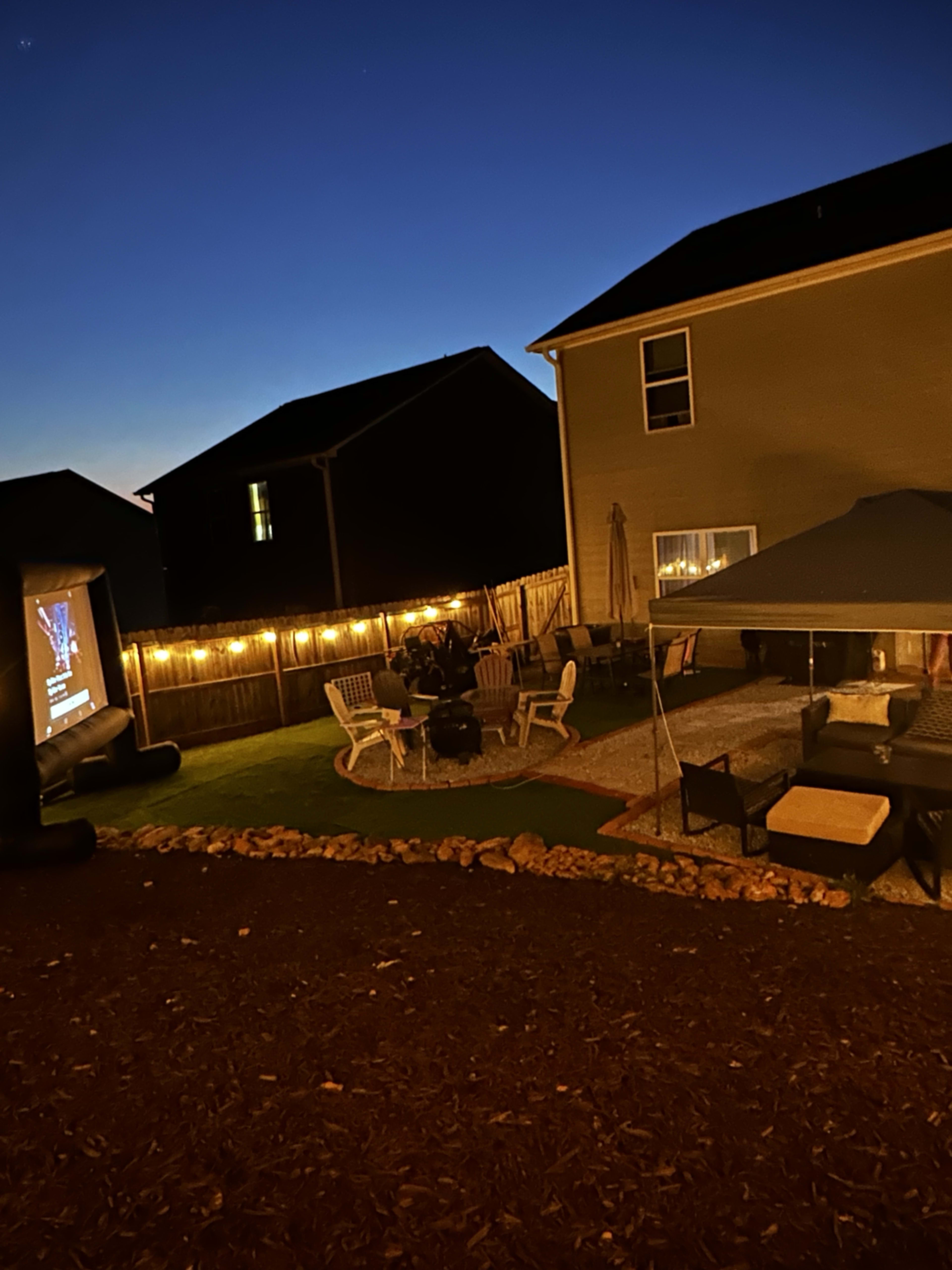 A backyard scene is illuminated by string lights with a projector showing an image on a screen, surrounded by seating areas and a patio.
