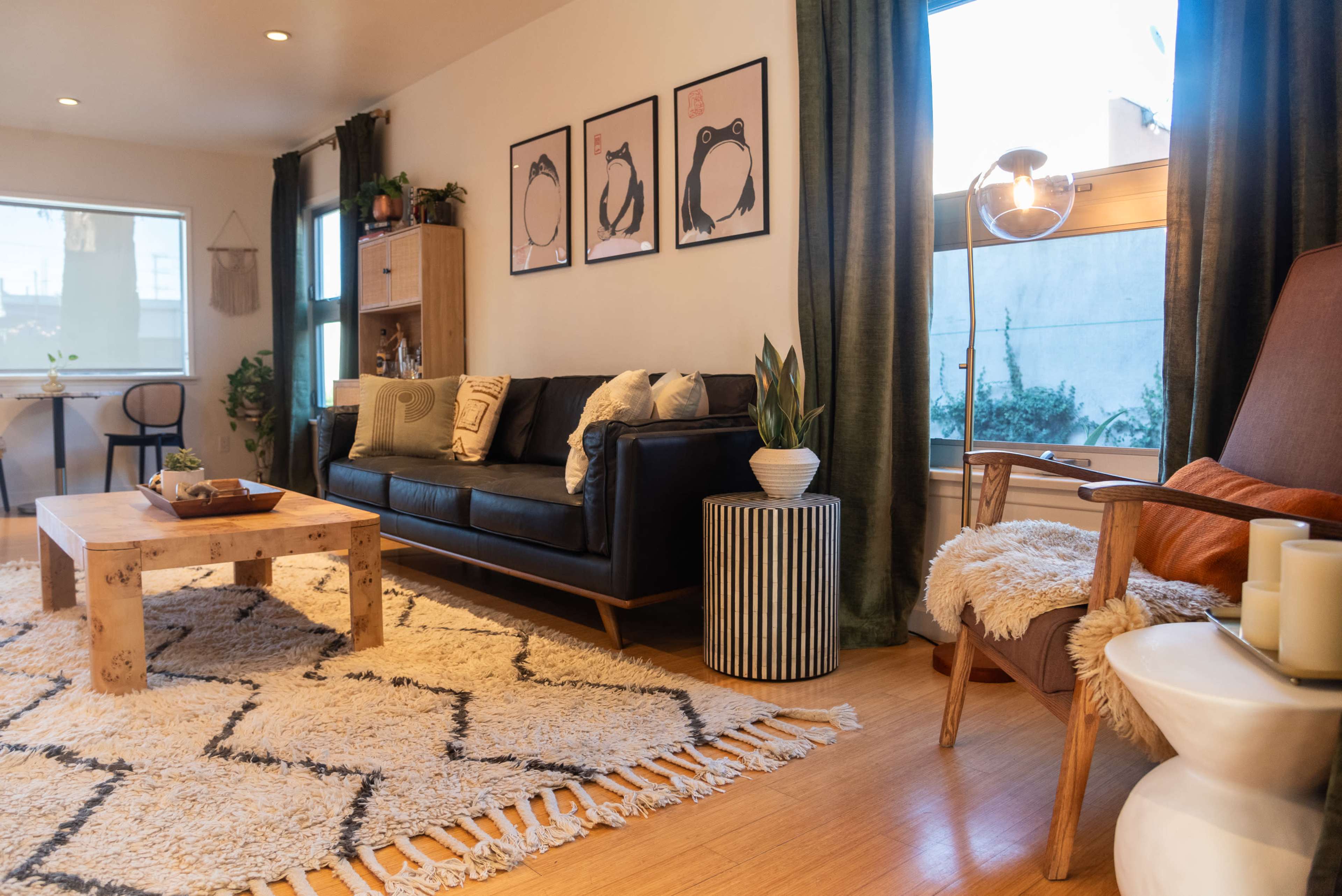 The image shows a modern living room featuring a dark sofa, a wooden coffee table, and art prints on the walls, with a rug covering the floor and plants in various corners.