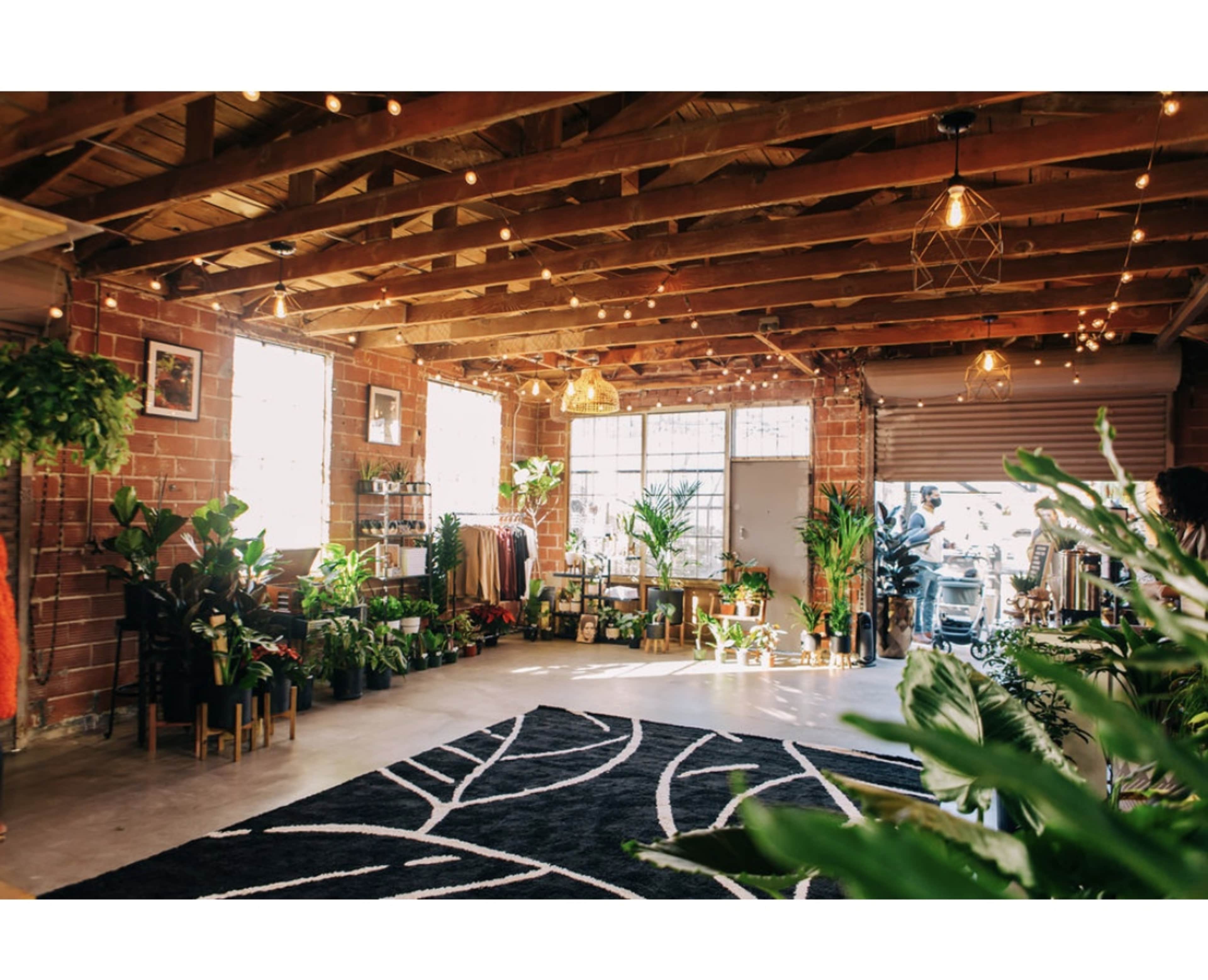 A spacious shop with exposed brick walls, wooden beams, and various plants arranged throughout the room, alongside clothing racks and decorative lighting.