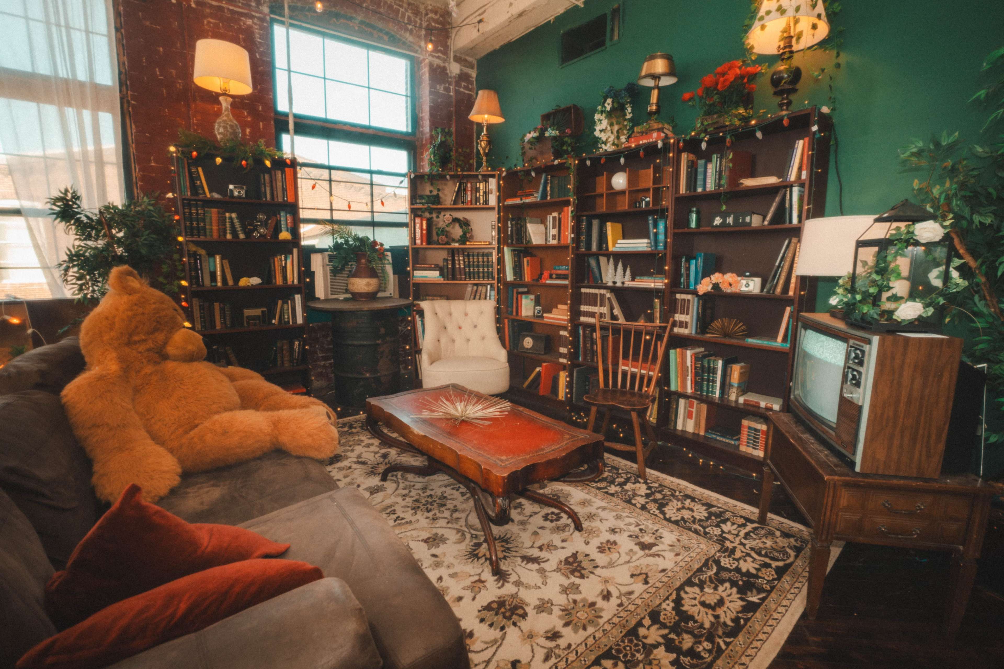 A cozy reading nook features a large stuffed bear, a wooden coffee table, bookshelves filled with books, and vintage furniture in a warm, well-lit room.