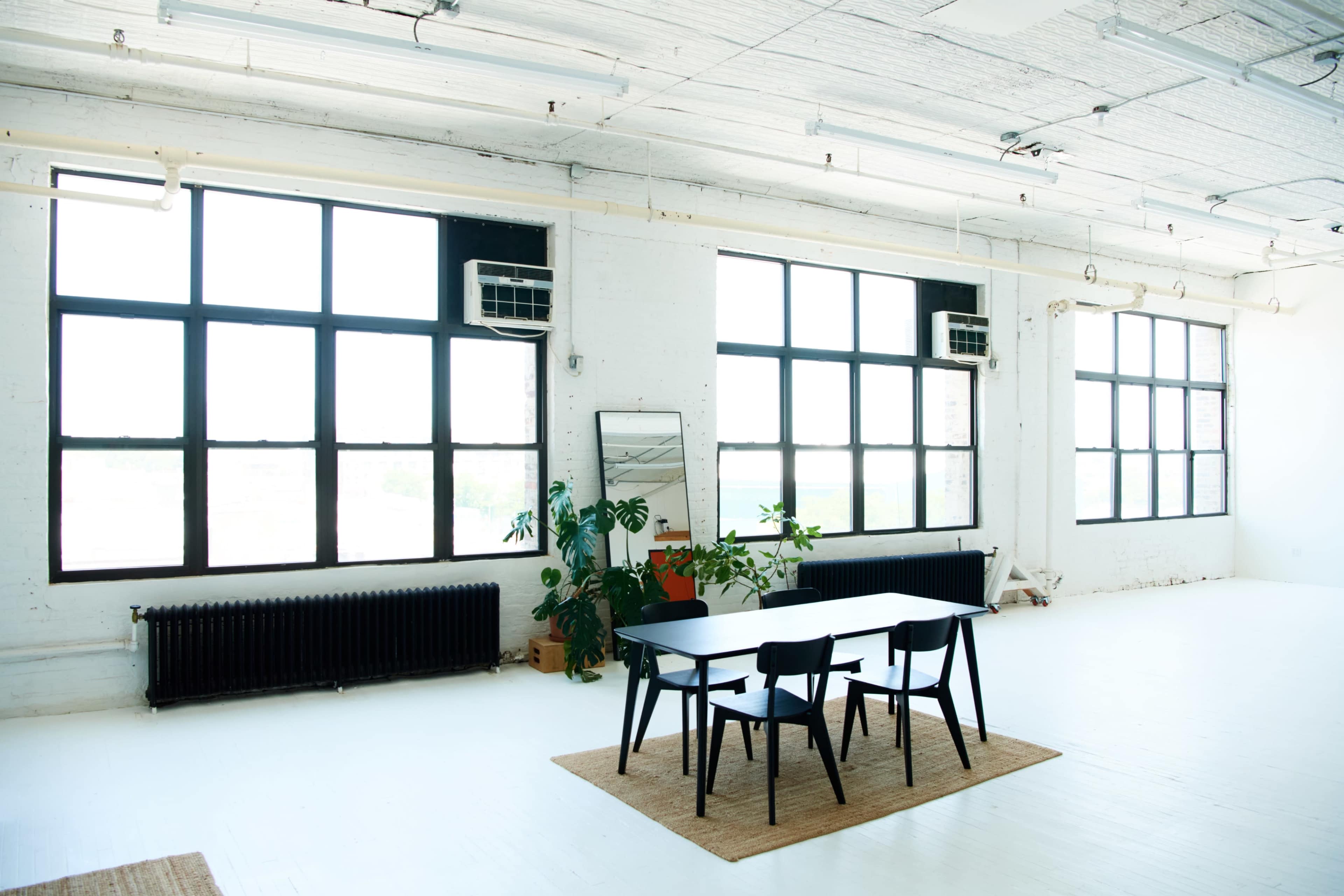 A spacious, well-lit room features a black table with chairs, large windows, a mirror, and potted plants against a white floor.