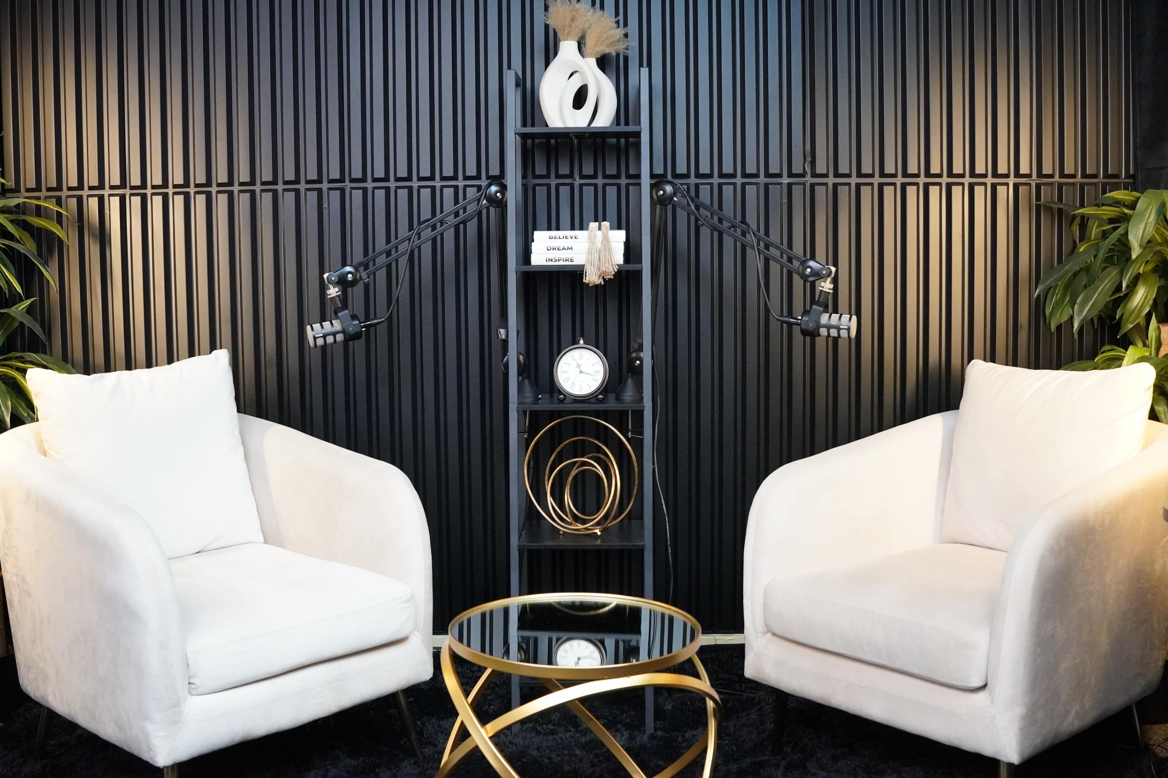 The image shows a modern interview setup featuring two white armchairs facing each other, with a small glass table in the center and a black wooden shelf displaying decorative items and a clock.