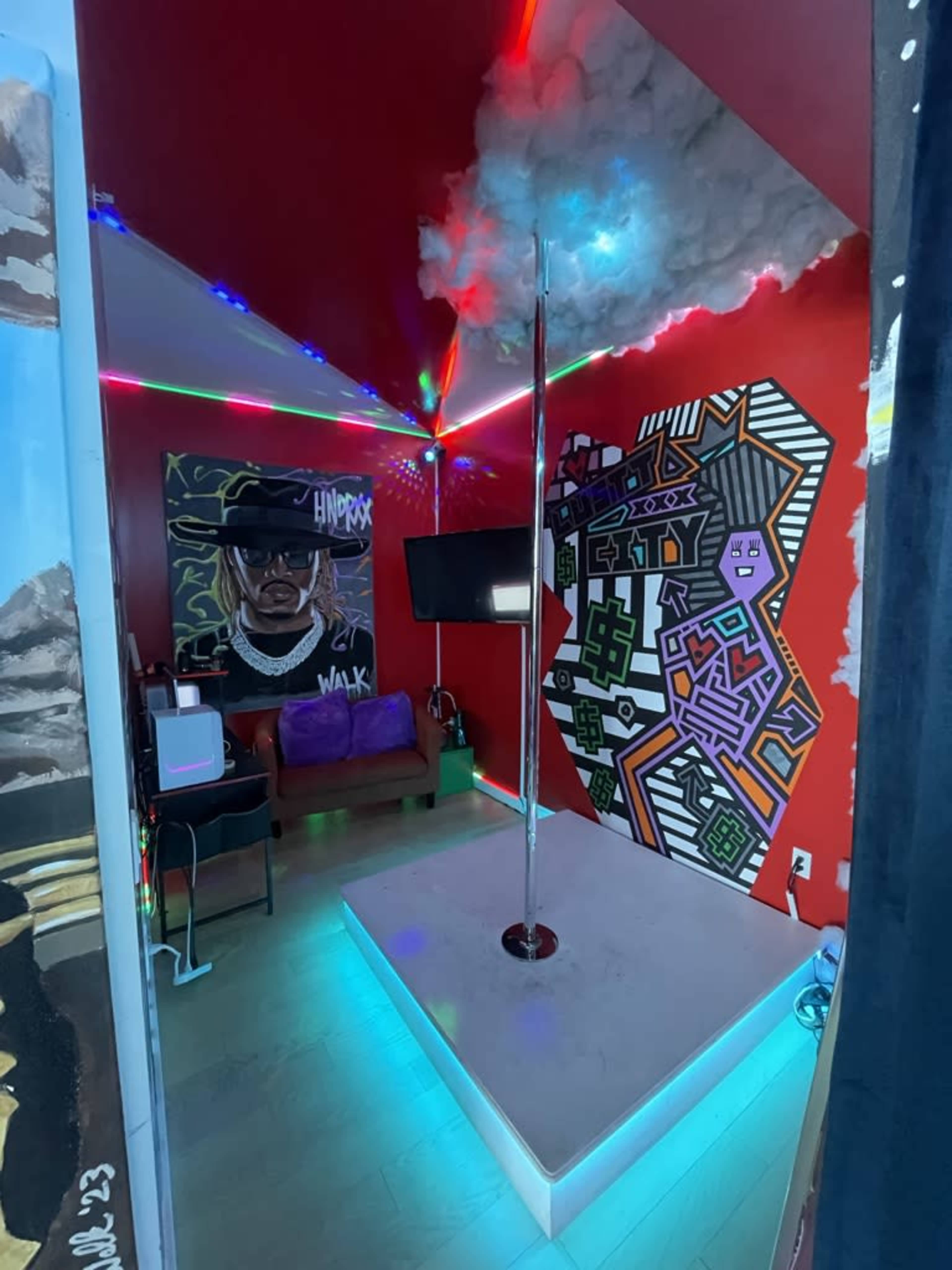 The image shows a dance studio with a mirrored floor, a pole in the center, colorful LED lights, and a vibrant mural on the red walls.