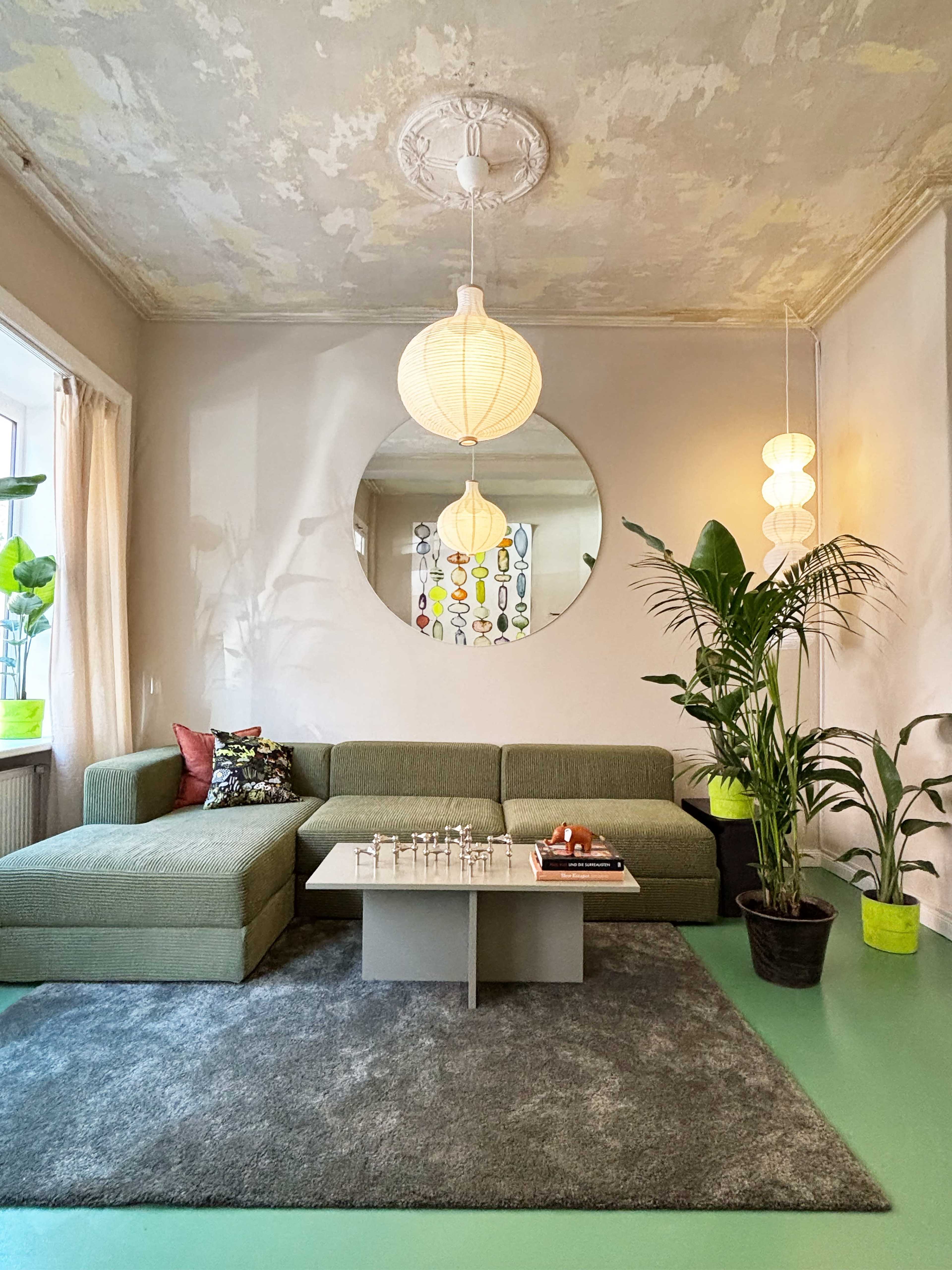The image shows a cozy living room featuring a green modular sofa, a round mirror, a coffee table with a chess set, and plants positioned by the window.