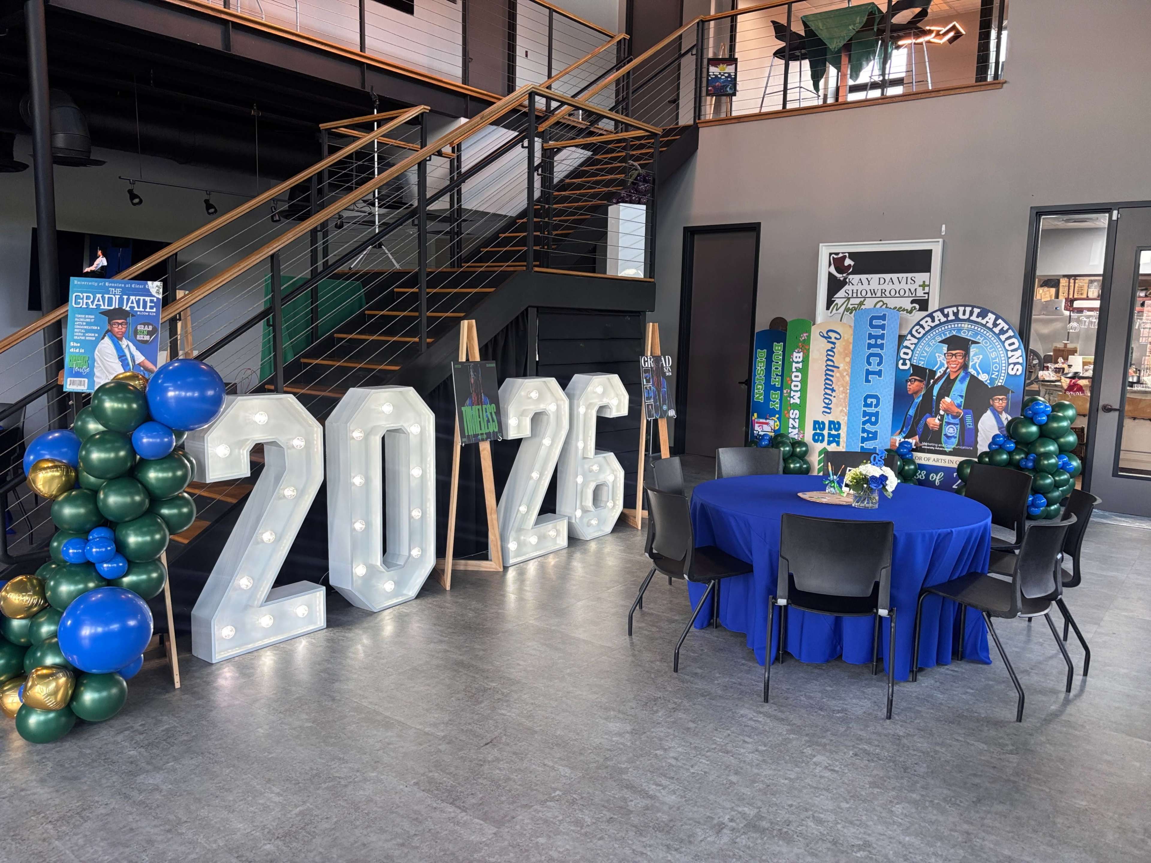 A decorated area features large illuminated numbers "2026," a table set with a blue tablecloth, and colorful balloons for a graduation celebration.