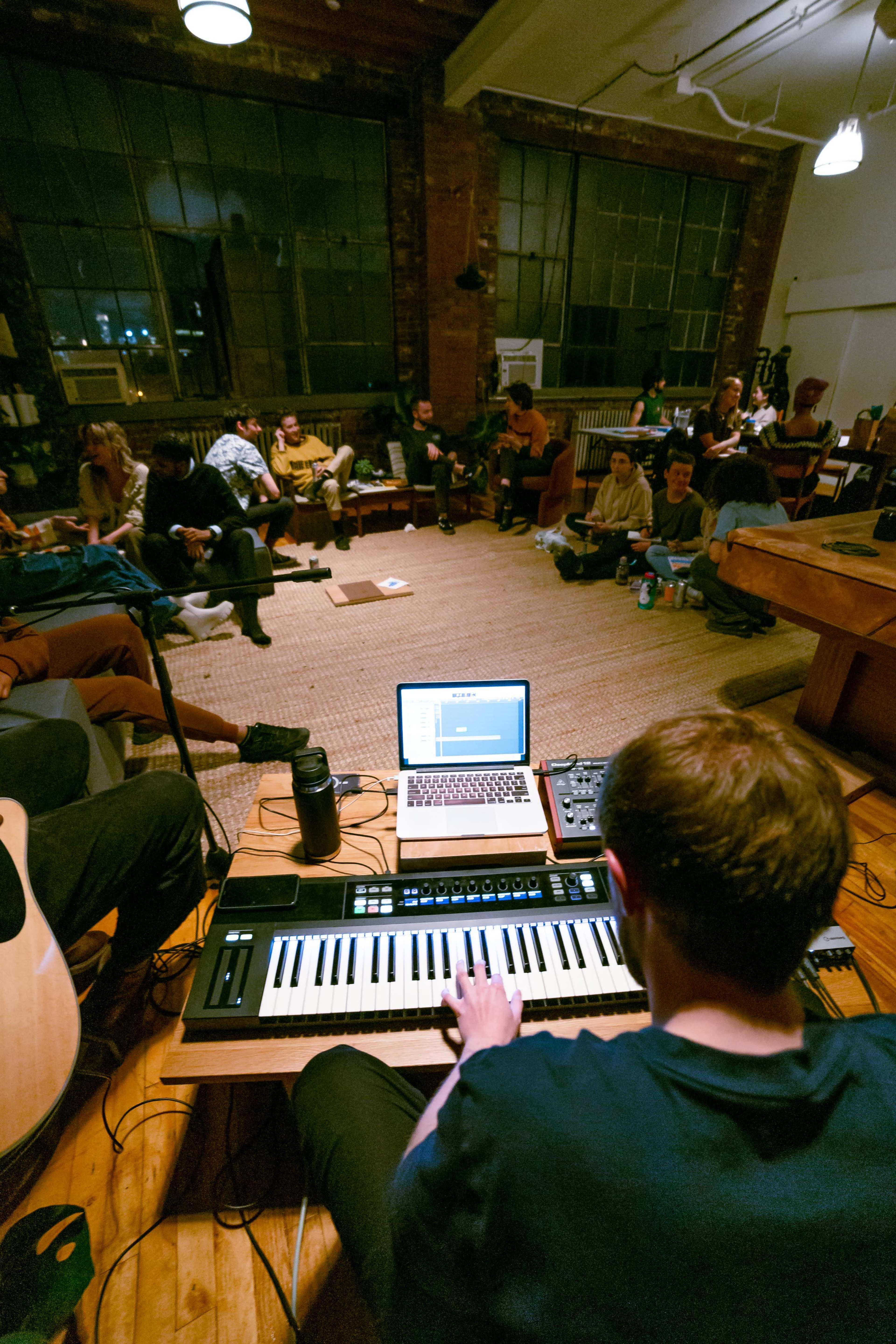 A group of people is gathered in a cozy room with large windows, while a musician plays a keyboard connected to a laptop on a table in the foreground.