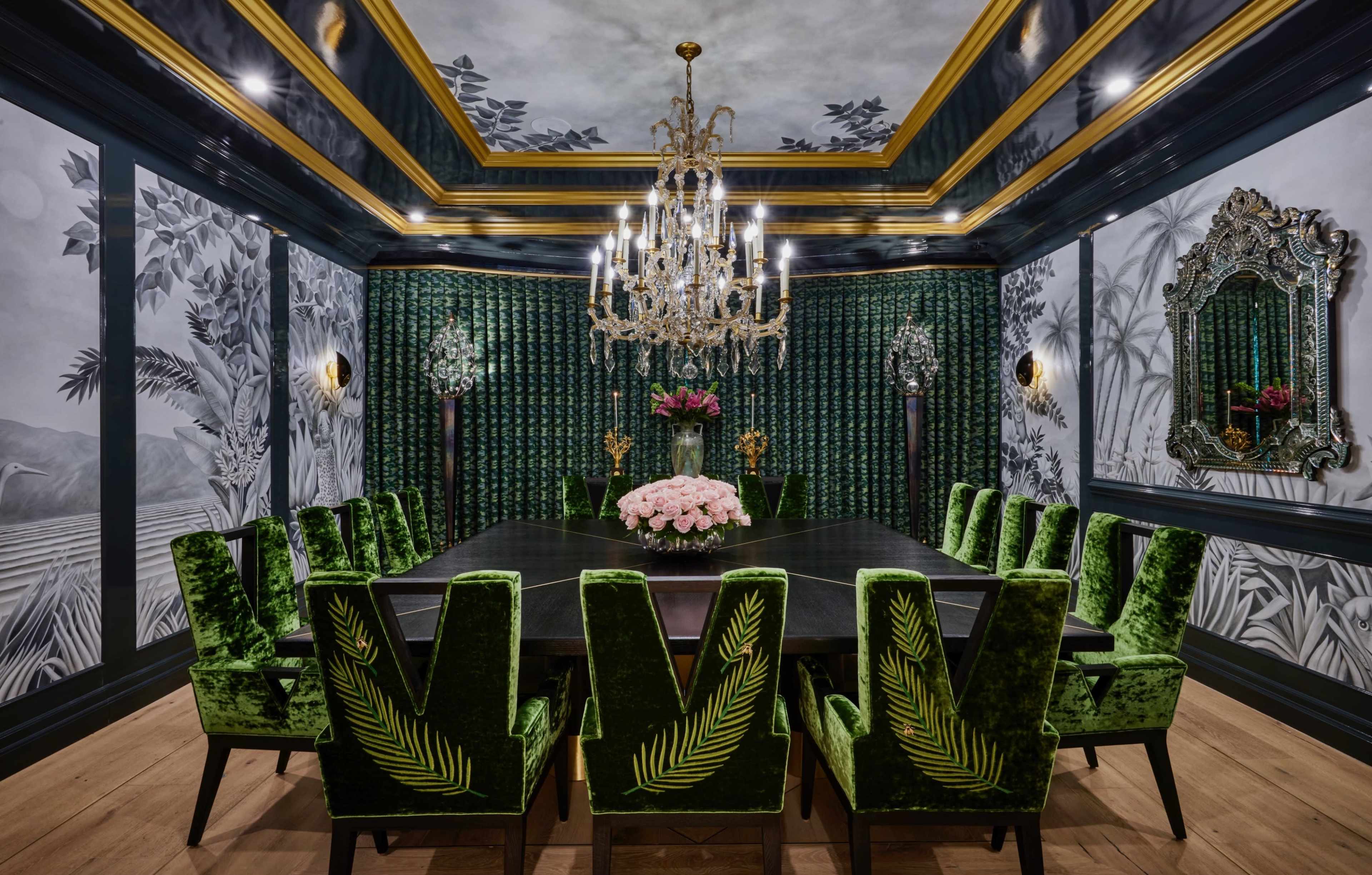 A long black dining table is surrounded by green velvet chairs, and the room features intricate wallpaper with tropical motifs and an ornate chandelier.