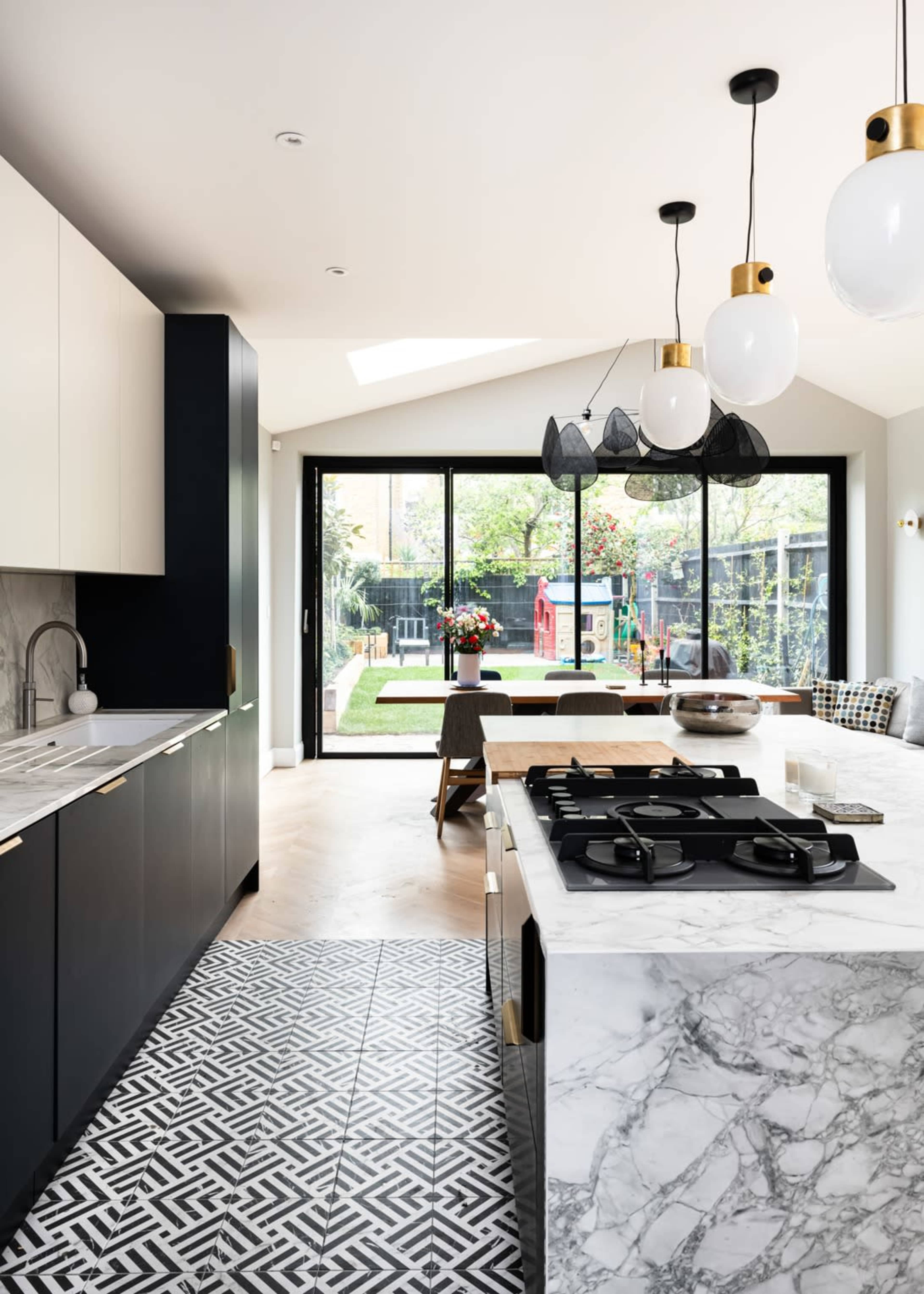 A modern kitchen features black cabinetry, marble countertops, and a geometric patterned floor, with large windows overlooking a grassy backyard.