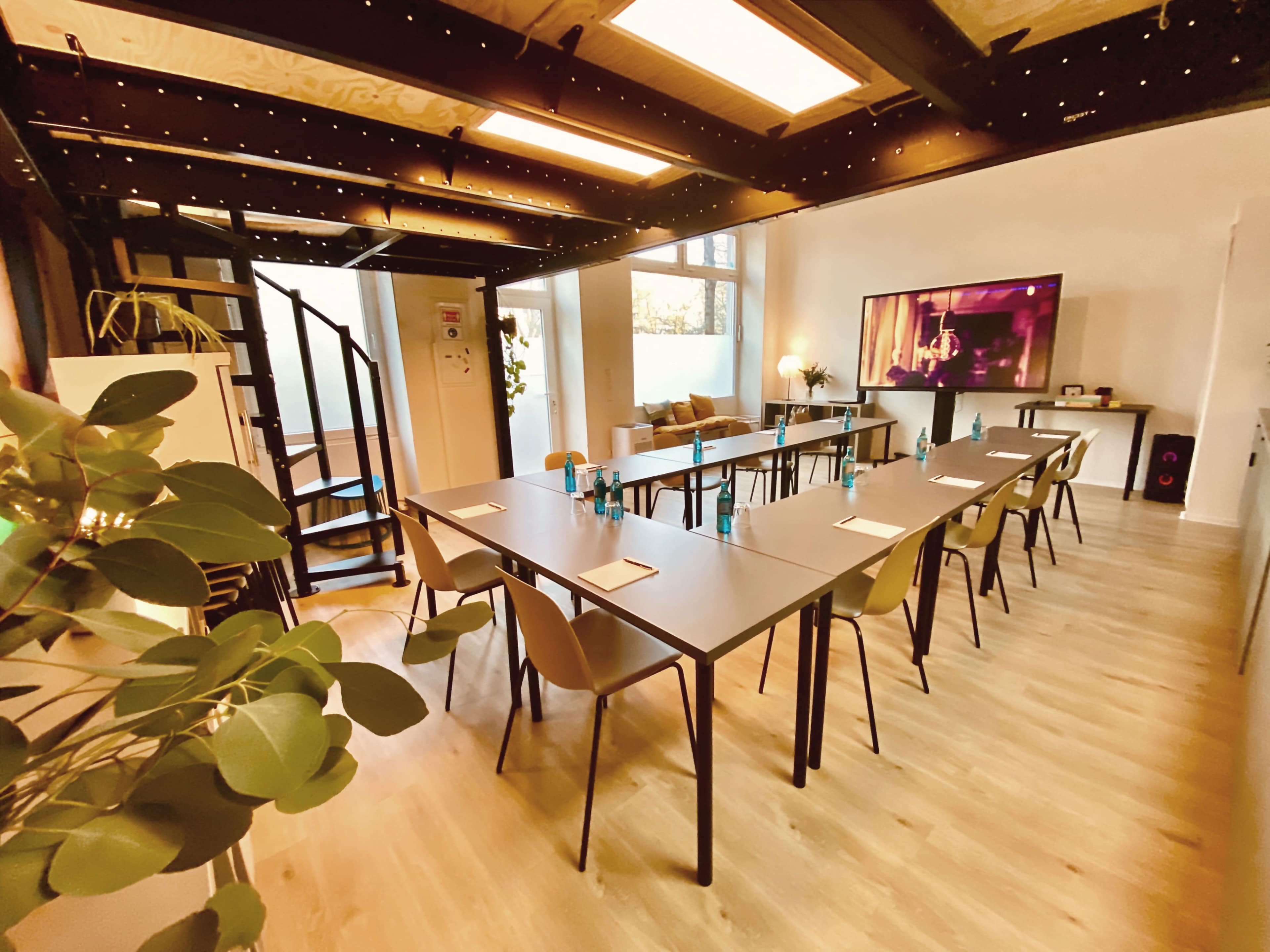 A modern meeting room features a set of long tables arranged for a conference, with chairs and a large screen at one end.