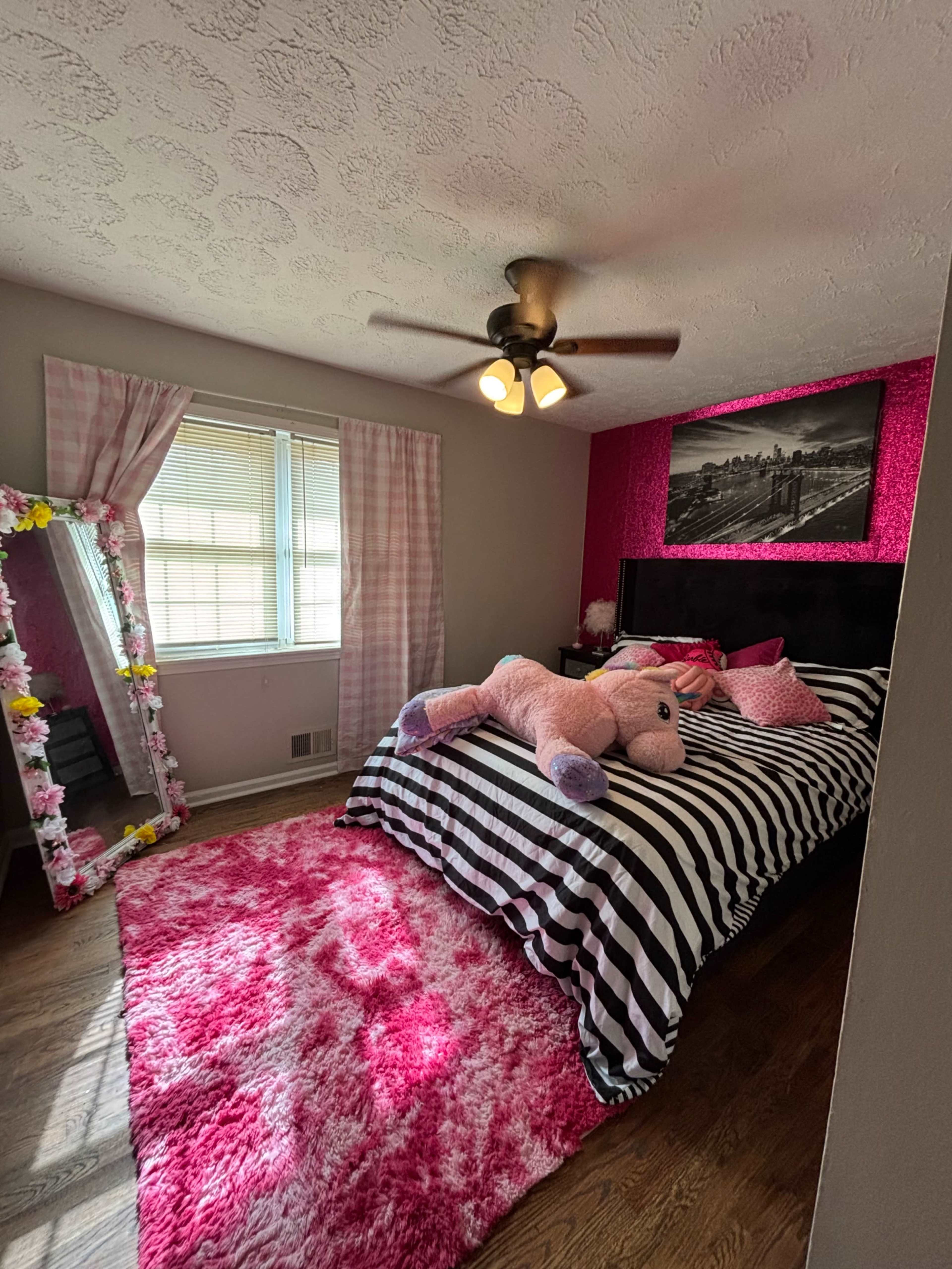 The image shows a bedroom featuring a striped bedspread, a large pink rug, a mirror adorned with flowers, and a plush unicorn toy on the bed.