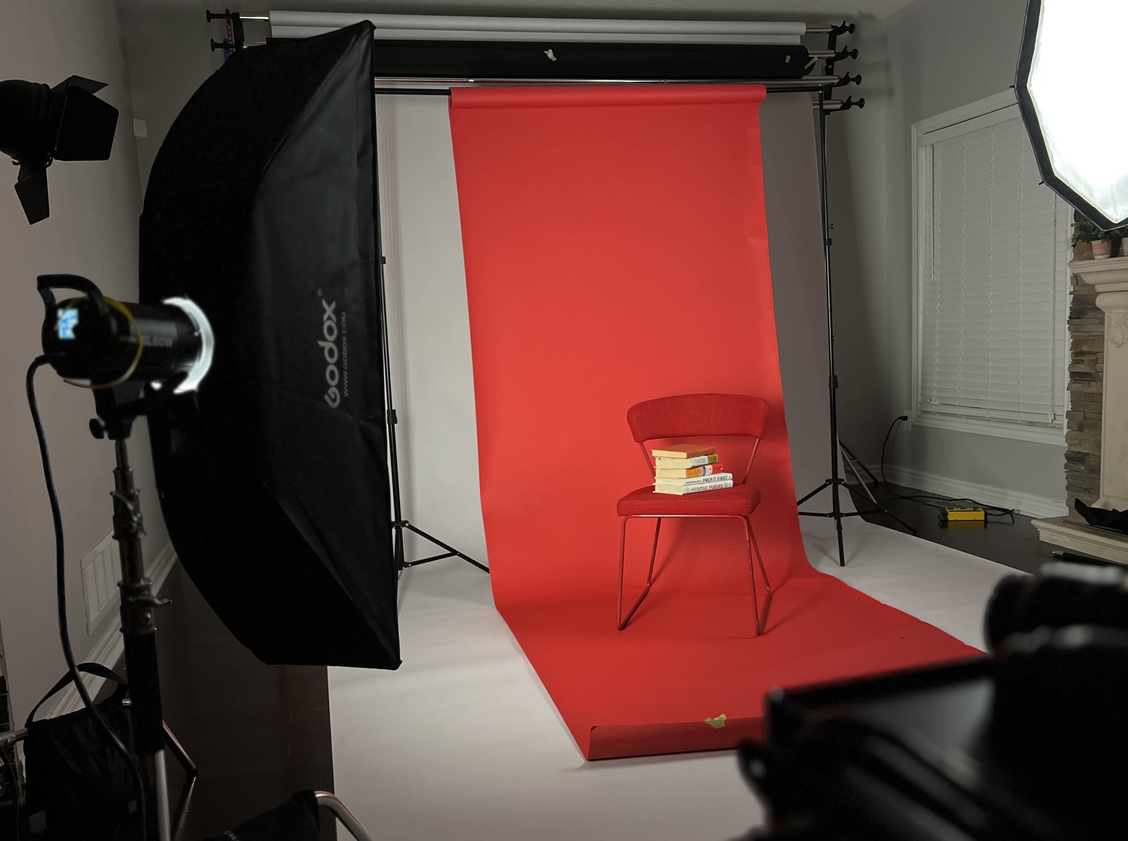 The image shows a red backdrop with a chair stacked with books, illuminated by studio lighting equipment.