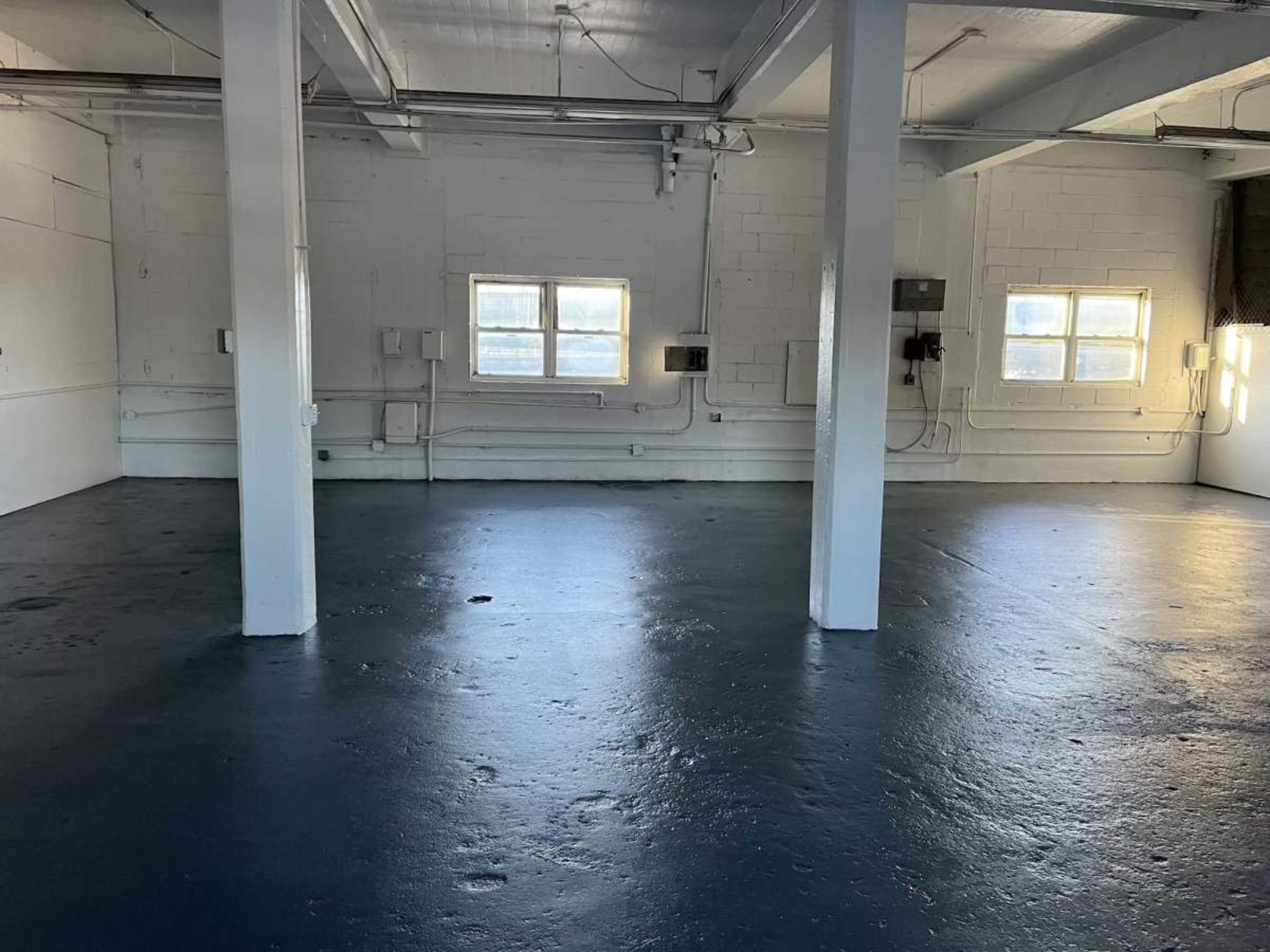The image shows a spacious, empty room with concrete flooring, white walls, and several windows.