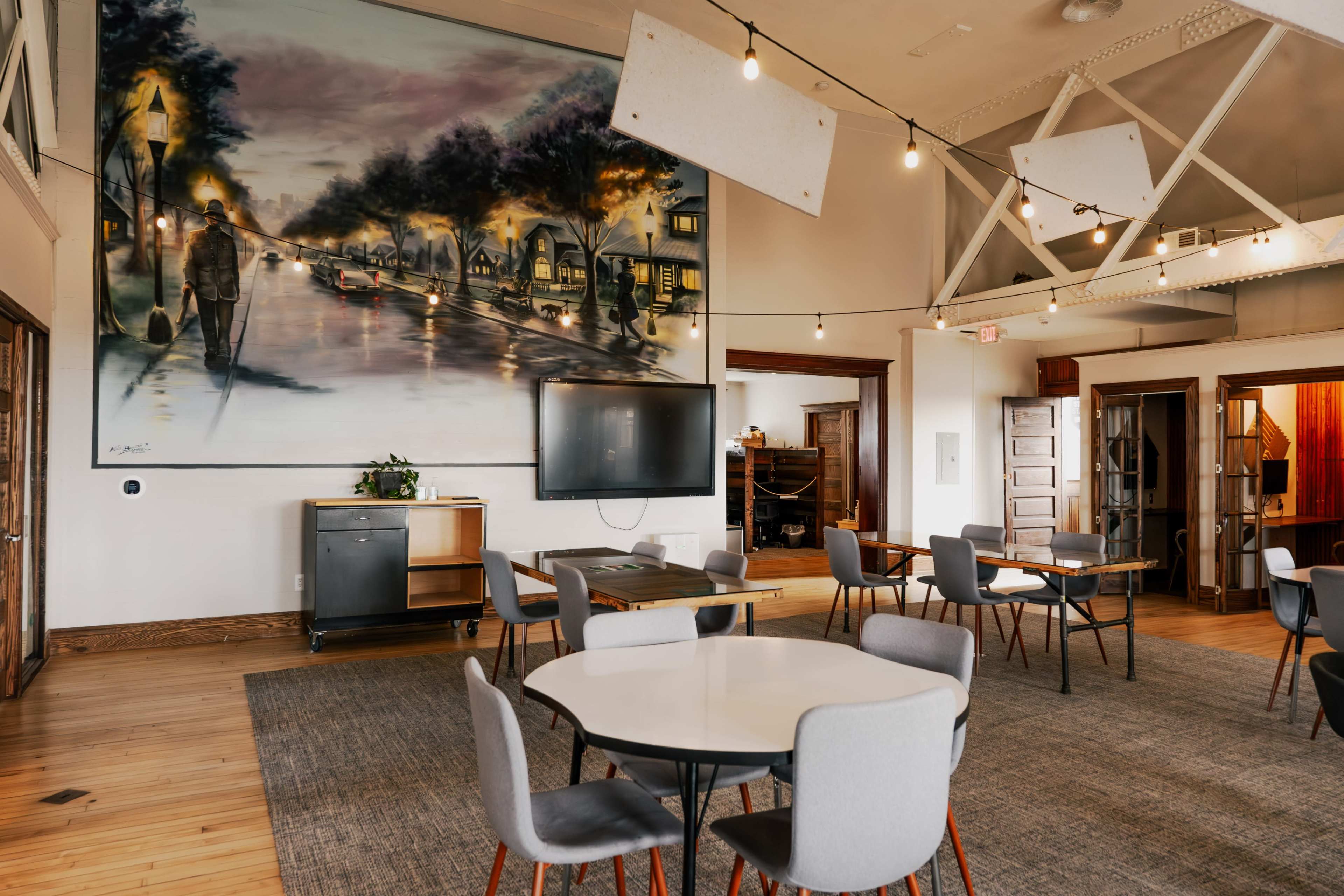 The room features a large mural depicting a street scene, with several round tables and chairs arranged on a wooden floor beneath hanging lights.