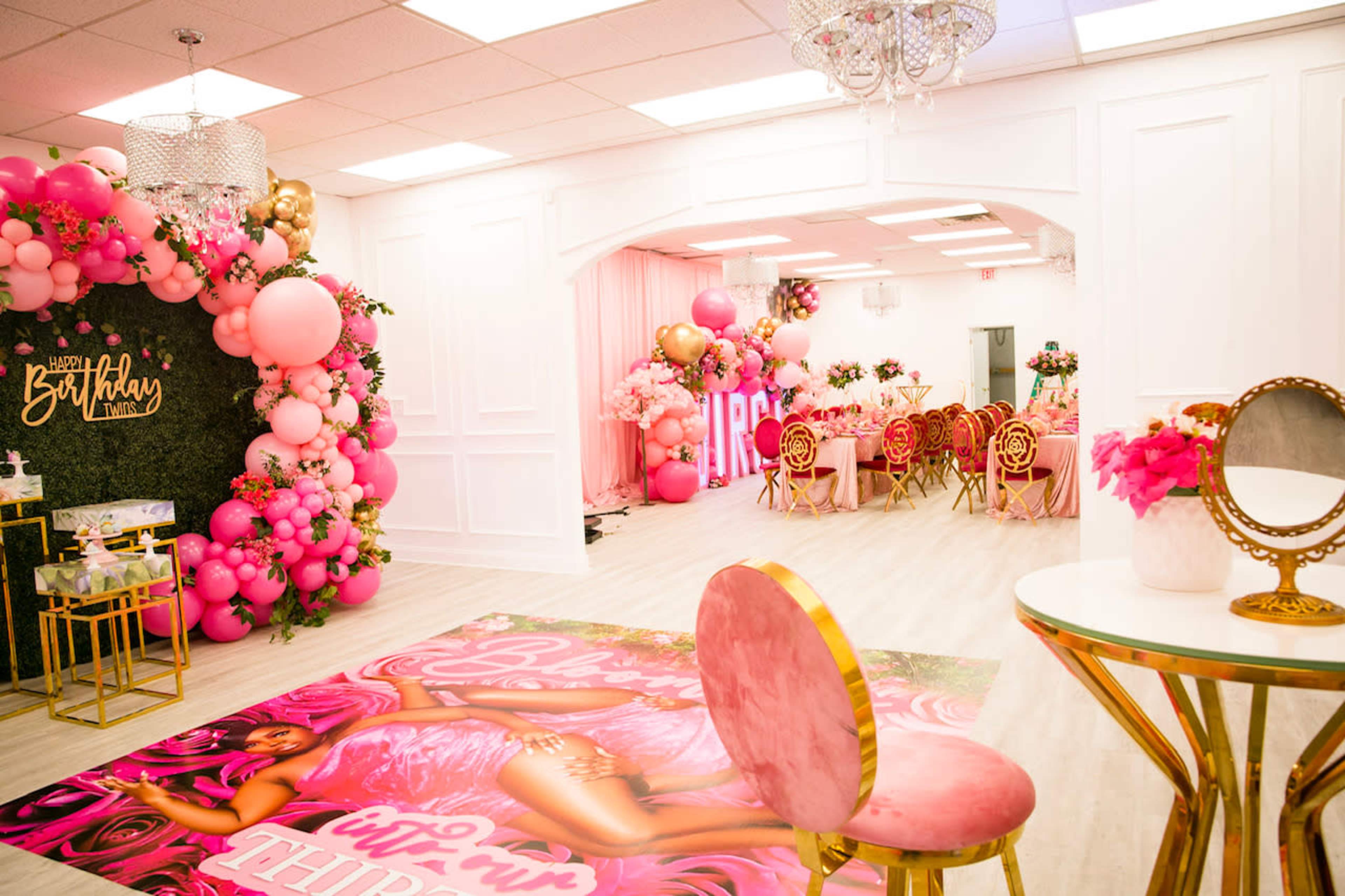 A decorated event space featuring pink balloon arrangements, a lush floral backdrop, and a themed floor mat with a seating area of gold chairs and a table.