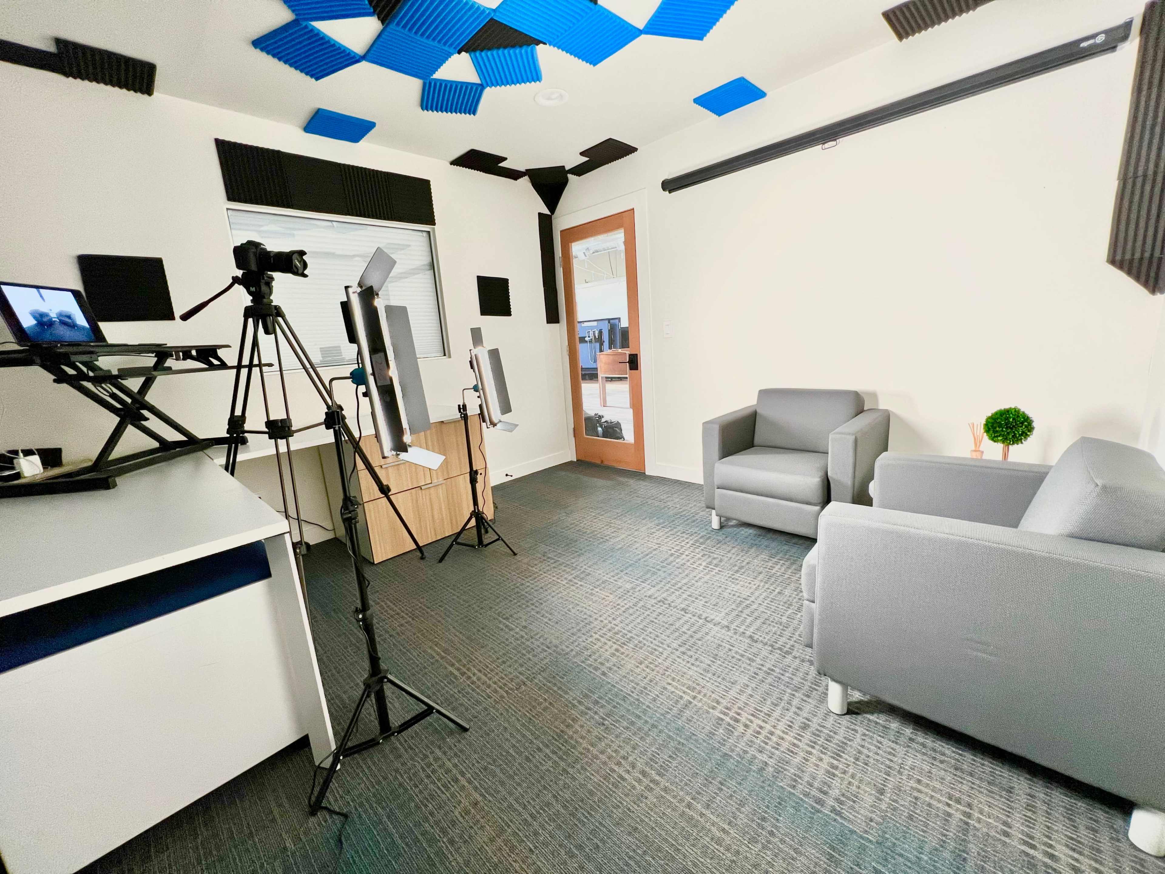A recording studio with two gray chairs, a desk with a camera setup, and several lighting stands positioned around the room.