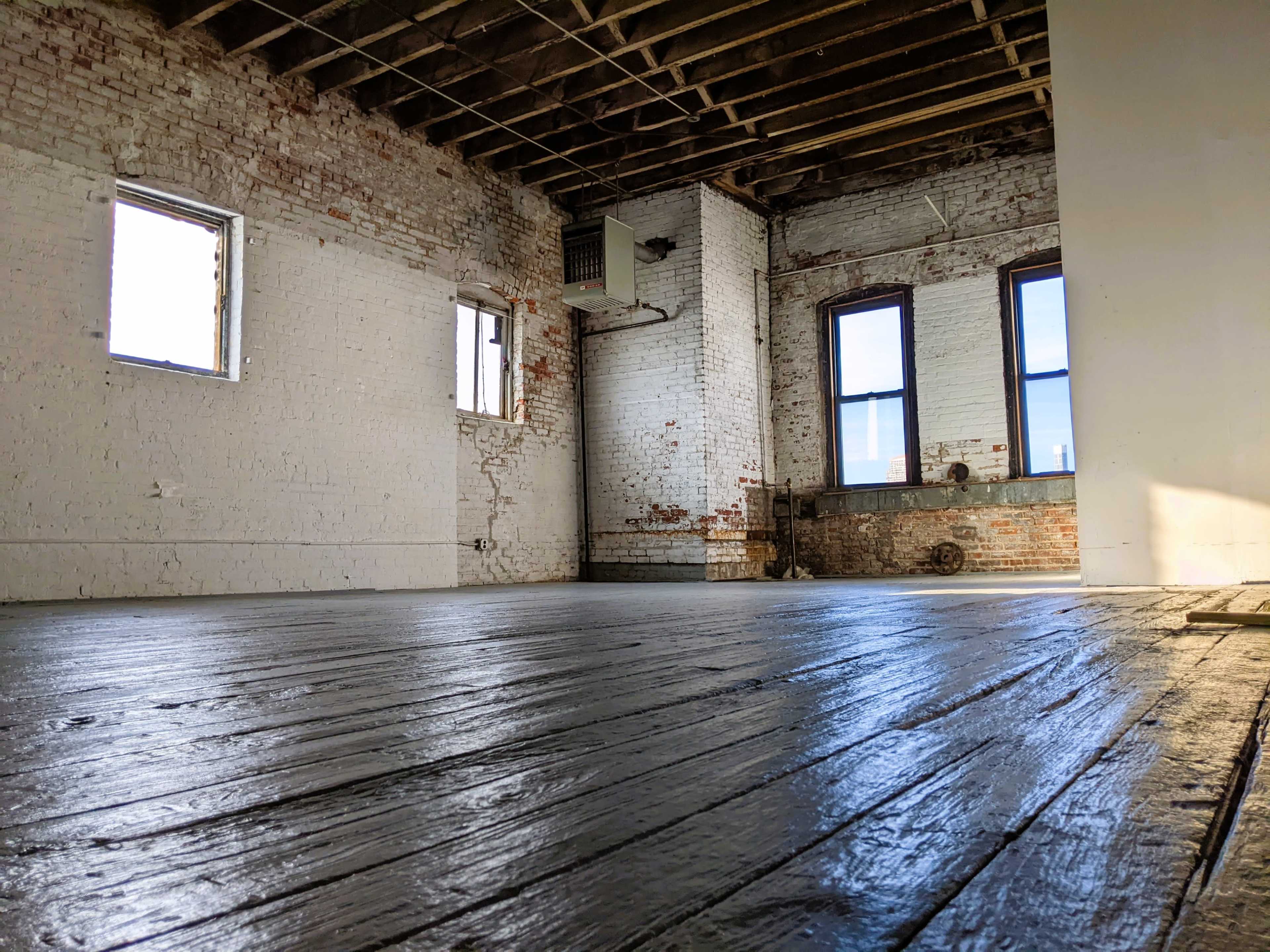 A vacant, industrial-style room with exposed brick walls, large windows, and a wooden floor reflecting light.