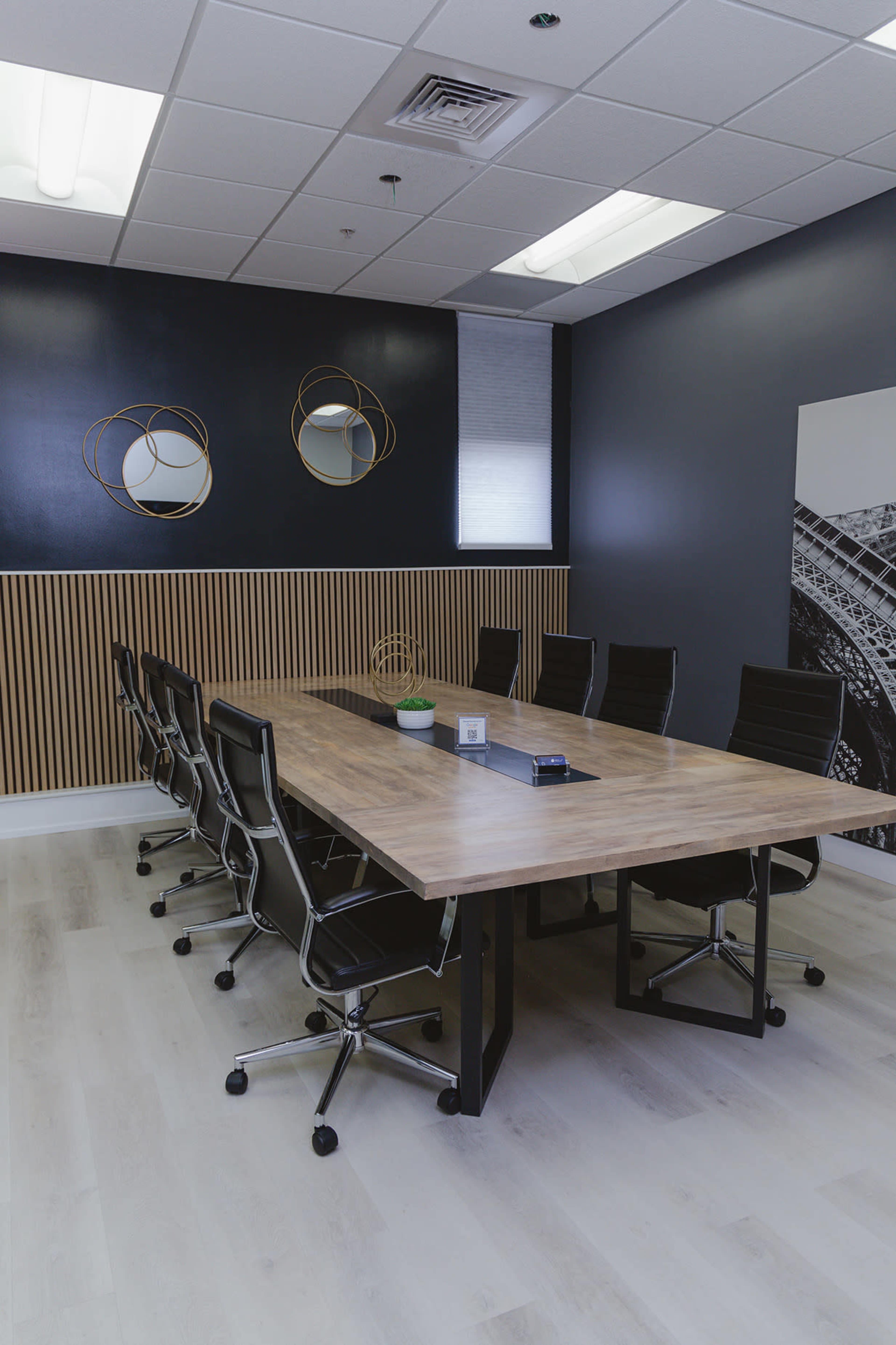 A modern conference room features a long wooden table surrounded by black rolling chairs, with decorative mirrors and a wall of wooden slats.
