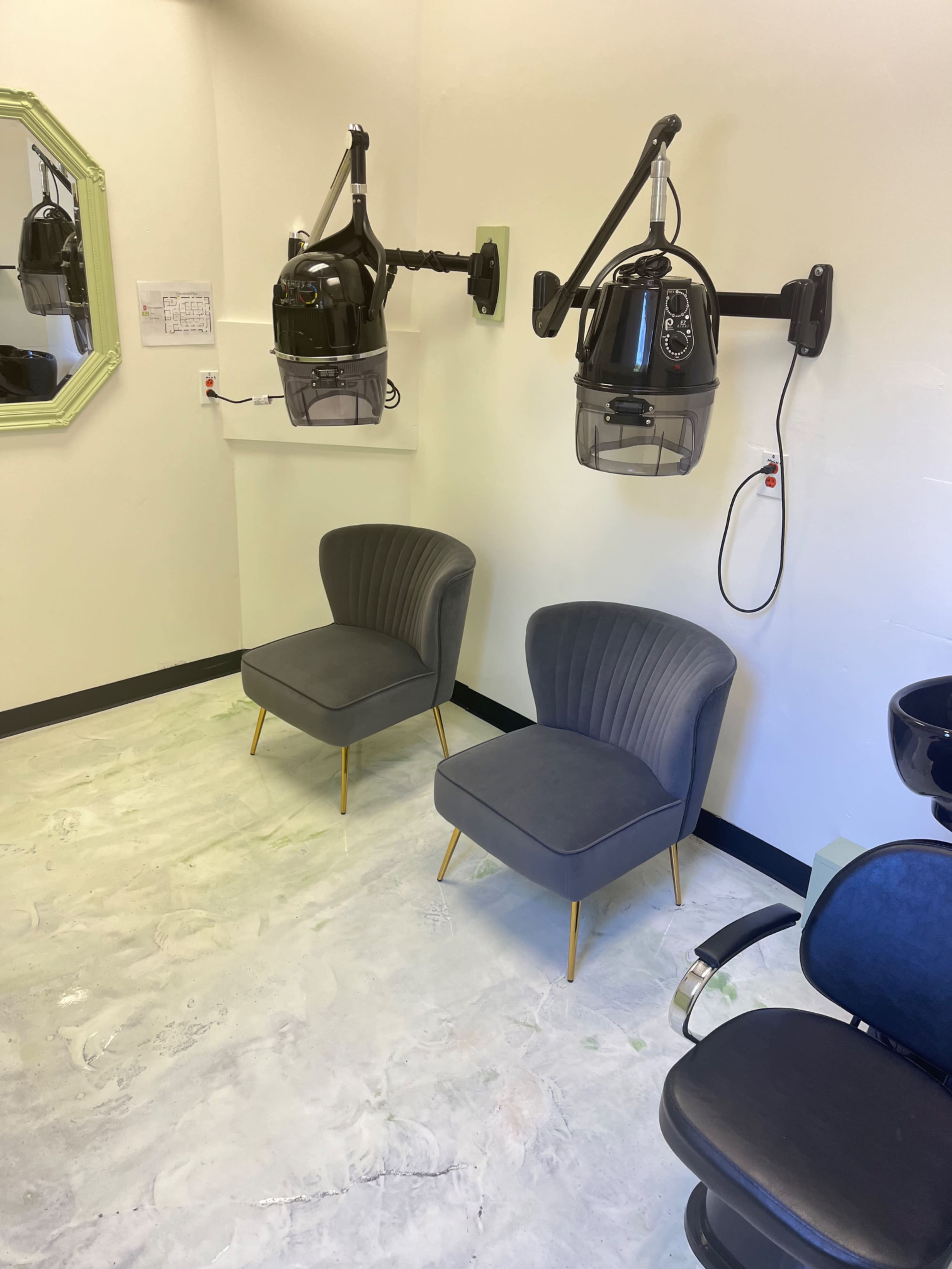 The image shows a hair salon interior with two hair dryers mounted on the wall, two gray upholstered chairs, and a black salon chair on a light-colored floor.