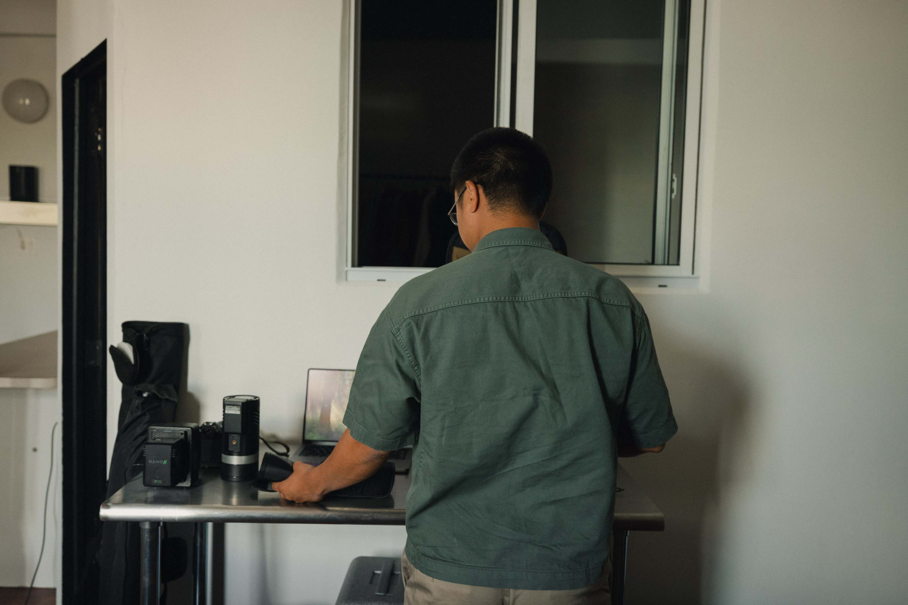 A person stands at a table with a laptop and camera equipment in a room with a window.