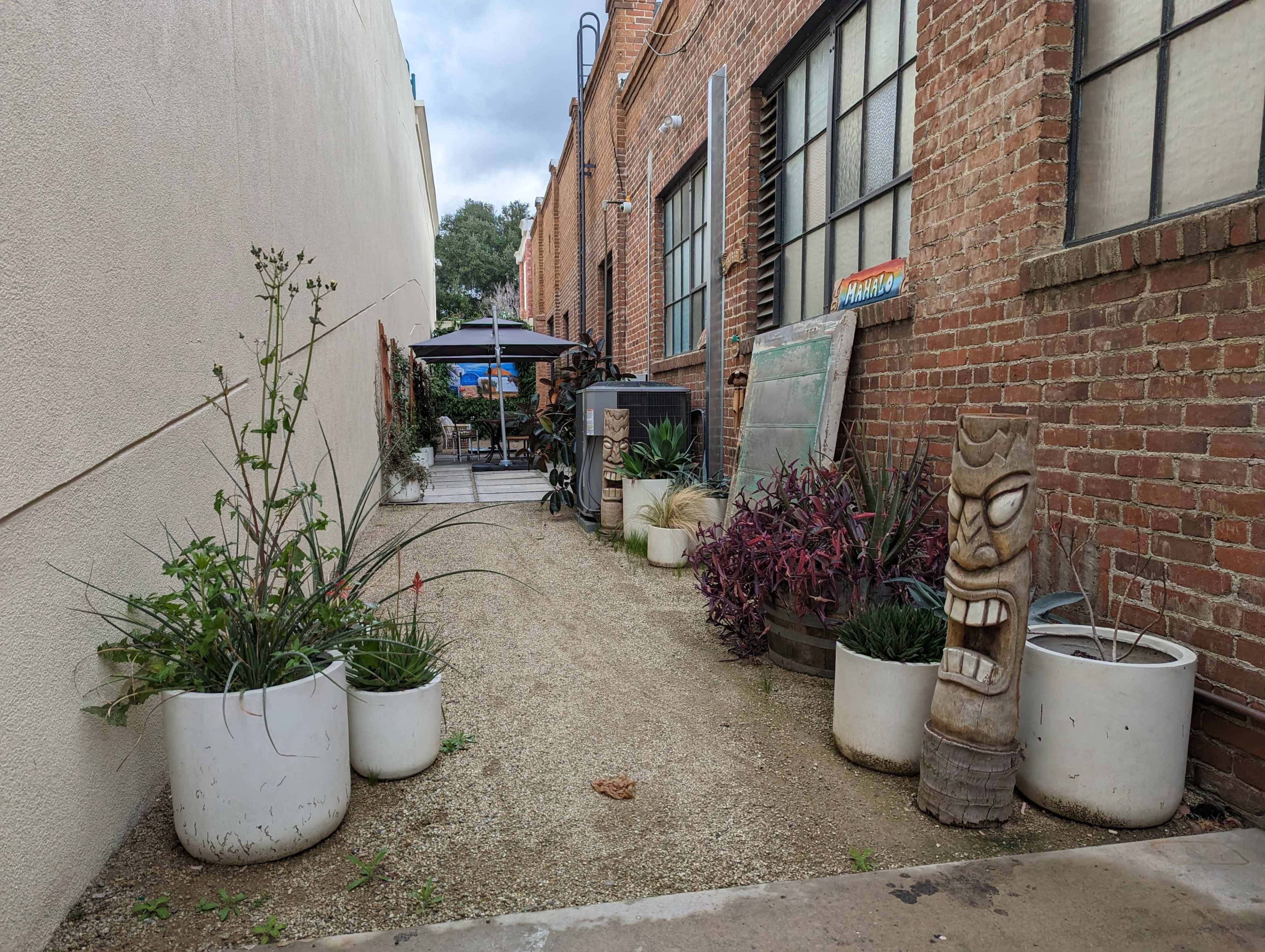The image shows a narrow alleyway between brick buildings, featuring potted plants and a wooden tiki sculpture along a gravel path.