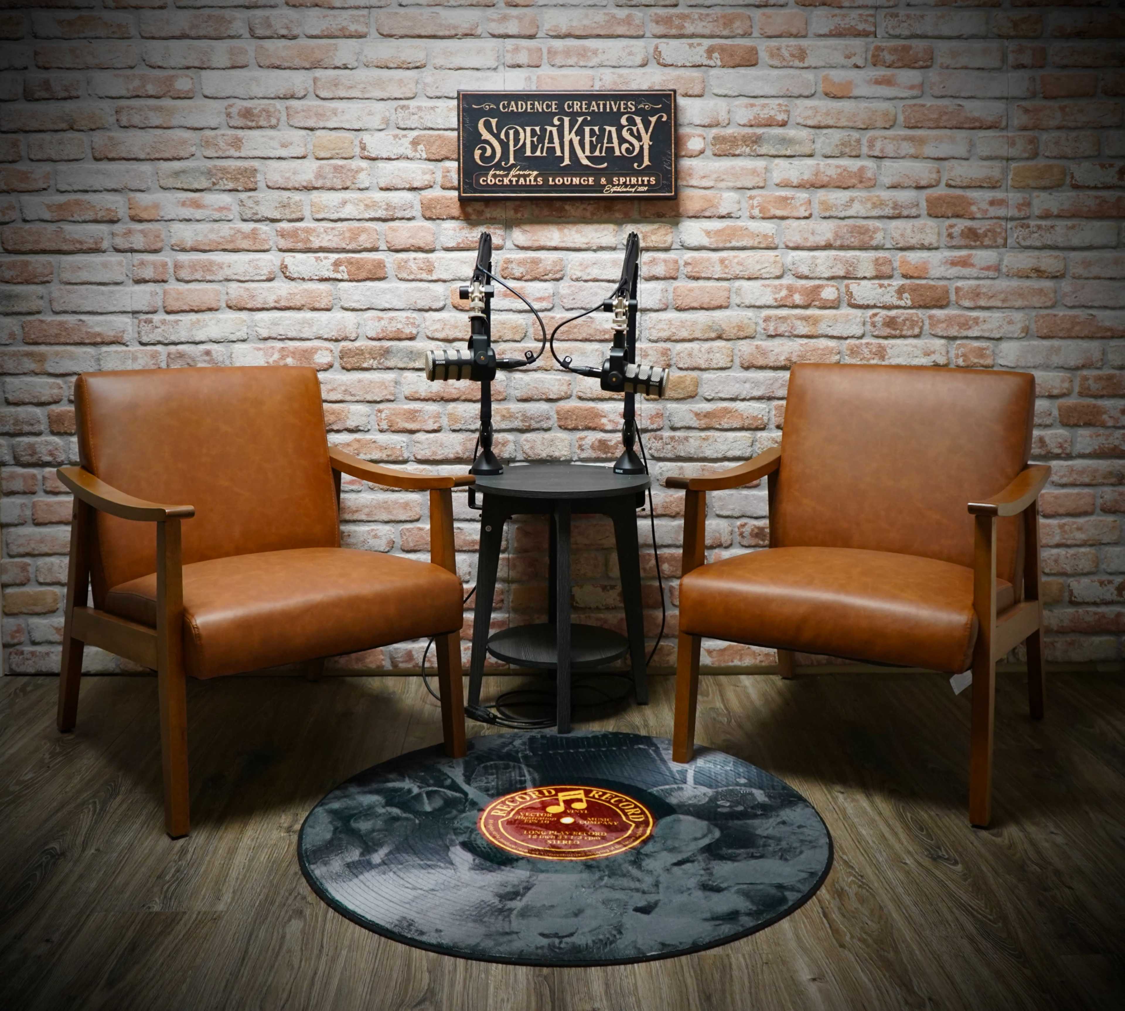 The image features two brown leather chairs placed opposite each other in a rustic room with a brick wall, a small round table, and two microphones on the table.
