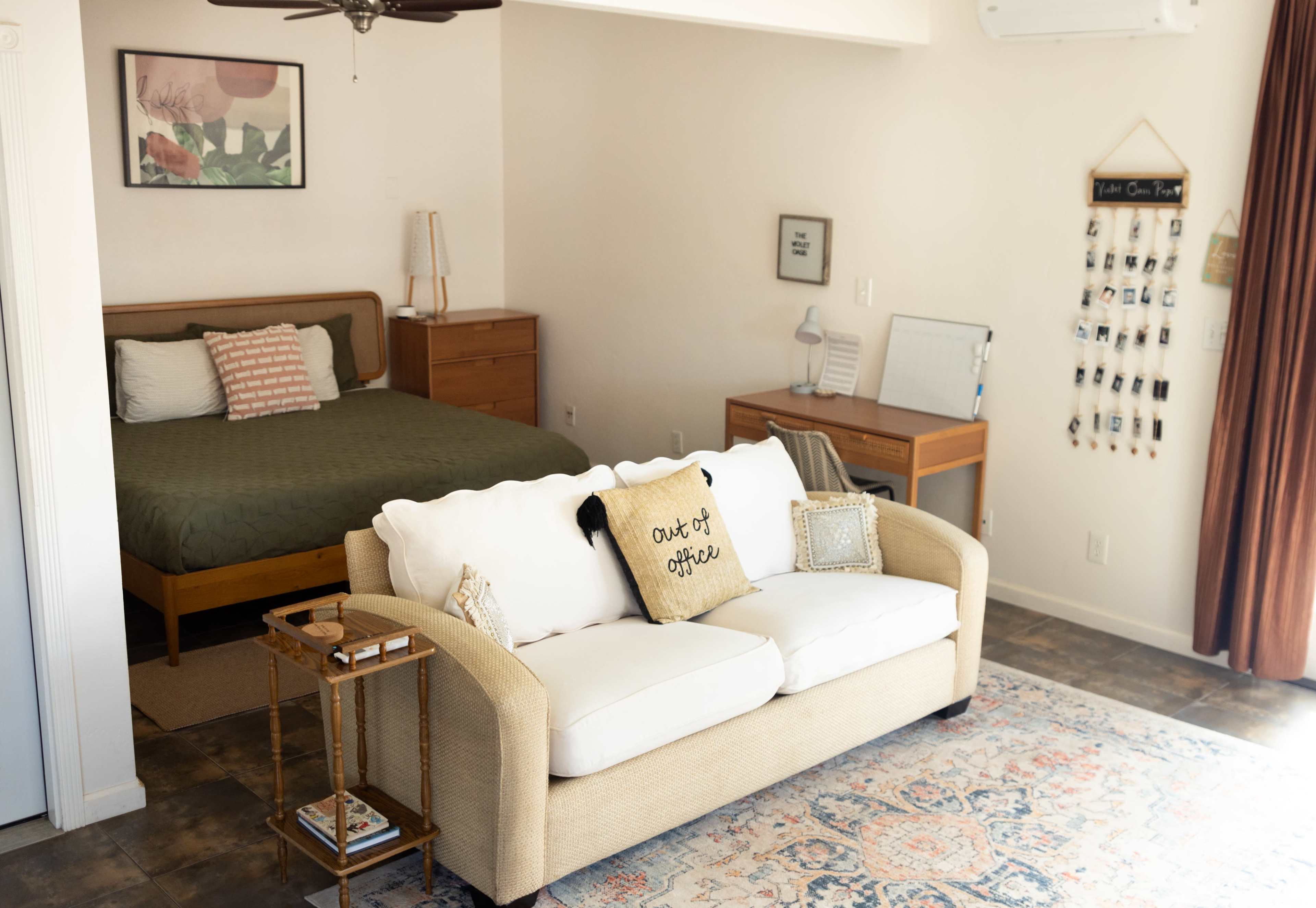 The image shows a cozy room featuring a beige couch with decorative pillows, a wooden bed in the background, and a small desk with a lamp.