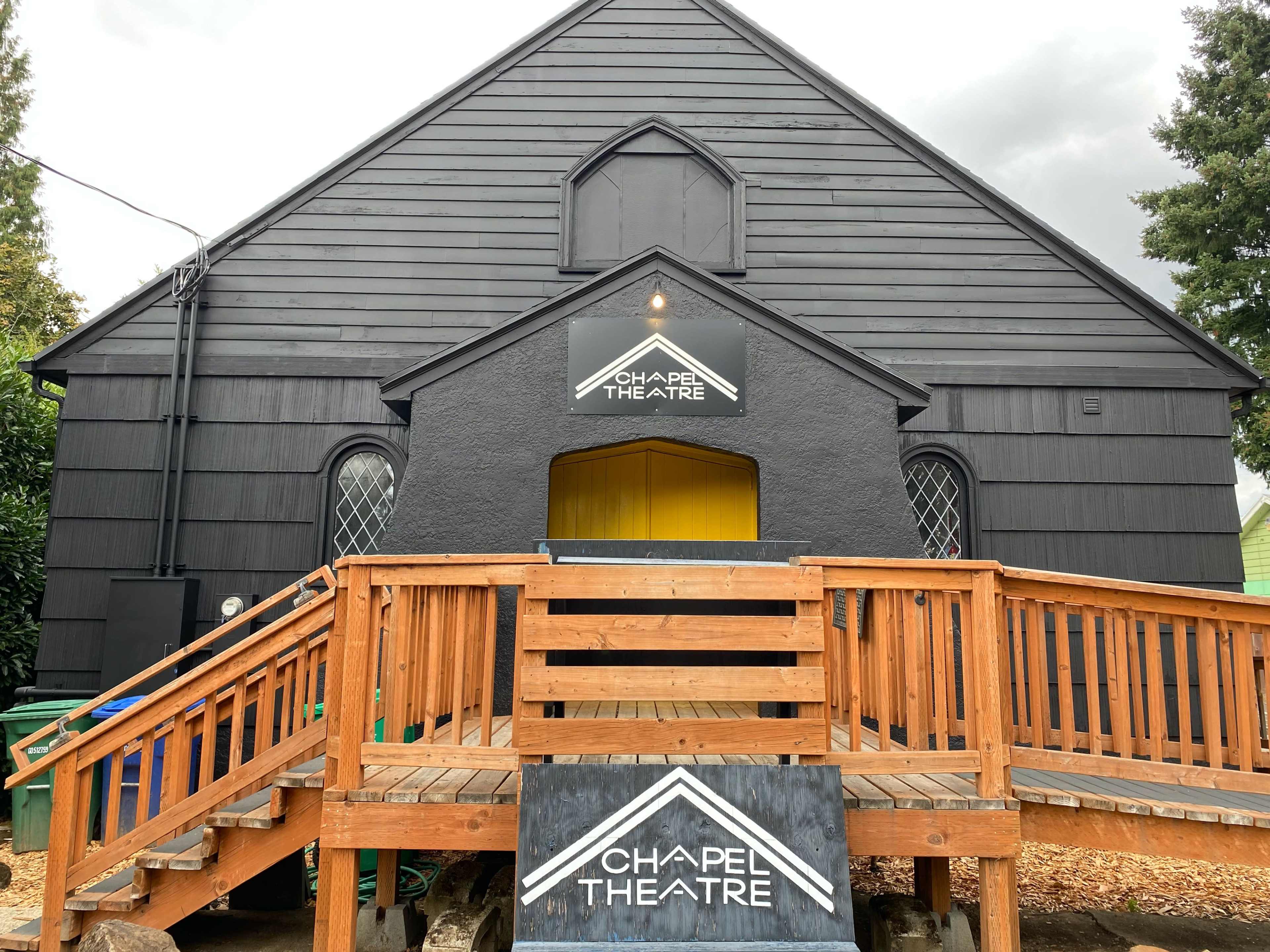 A black wooden theater building with a yellow entrance, featuring a wooden ramp leading to the front door and a sign that says "Chapel Theatre."