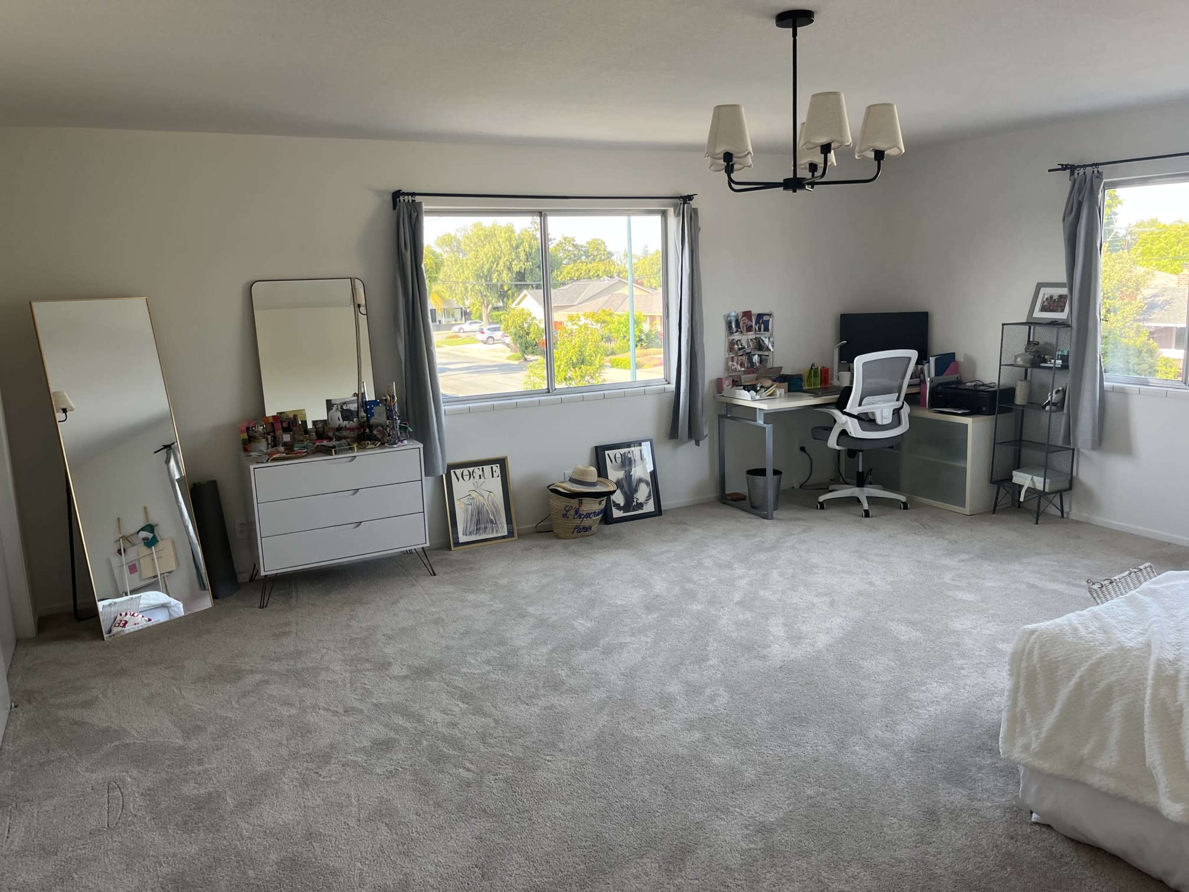 The image shows a spacious, well-lit room with a light carpet, a mirror, a dresser, and a desk with an office chair near two windows.