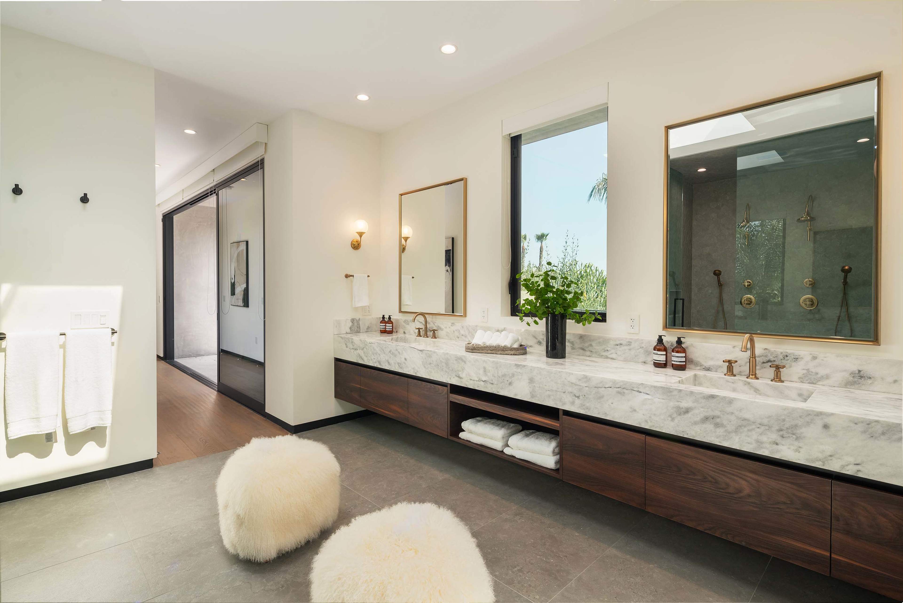 The bathroom features a double sink with a marble countertop, wooden cabinetry, two mirrors, and fluffy stools, with large windows allowing natural light and a view of greenery.