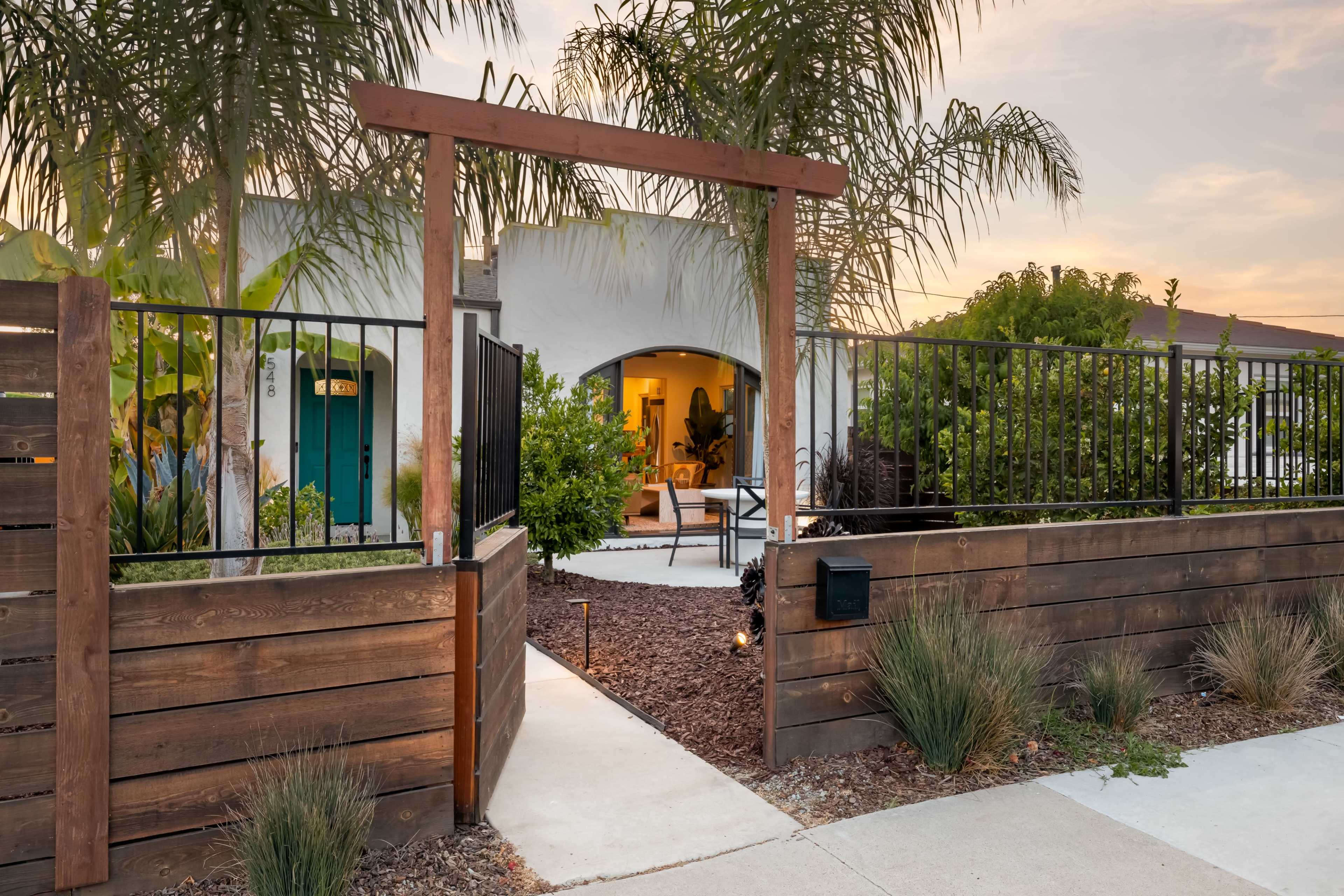 The image shows a wooden gate leading to a landscaped pathway that connects to a modern house surrounded by palm trees.