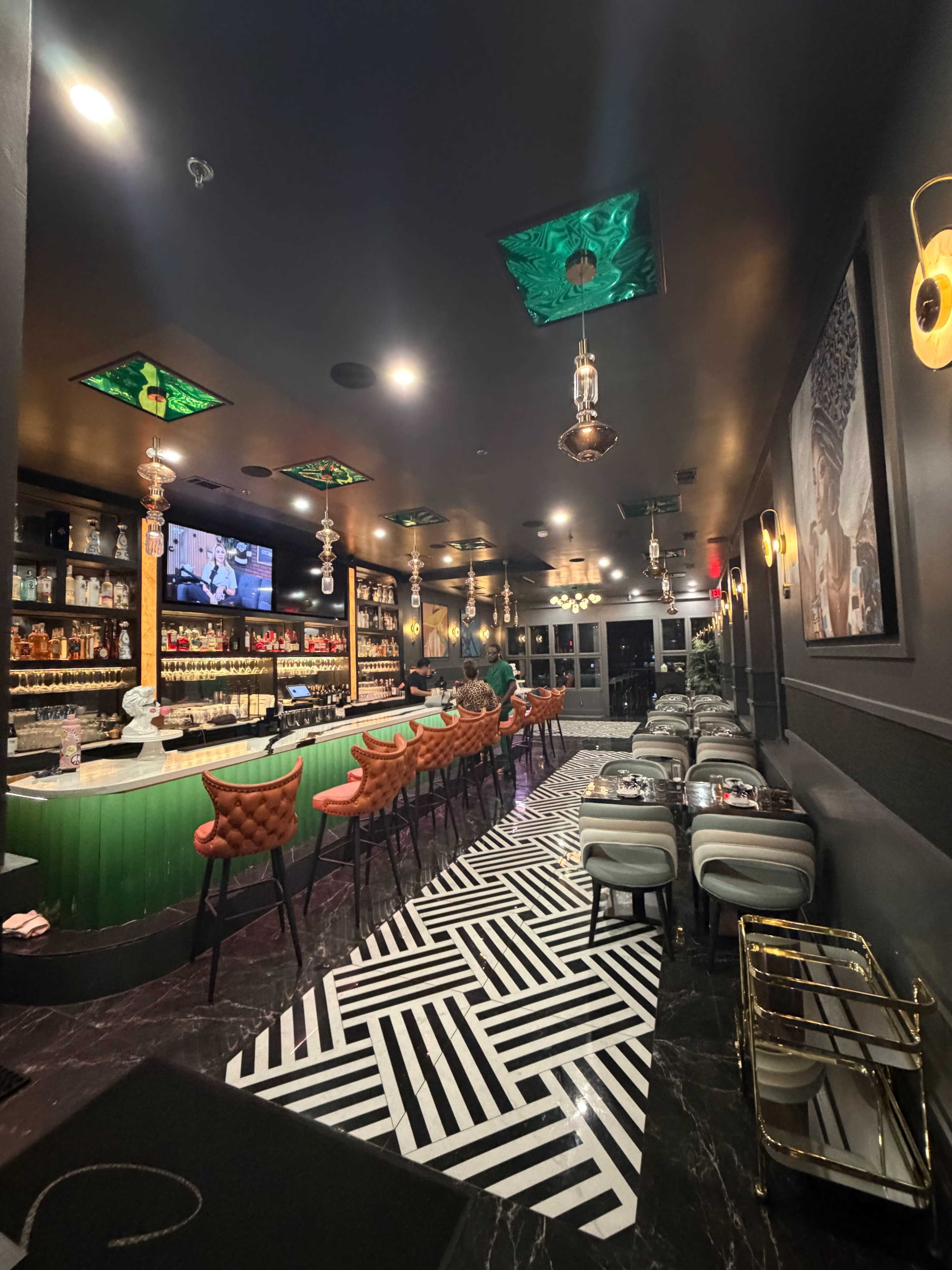 The image shows a modern bar interior with a green counter, patterned flooring, a variety of bottles on shelves, and stylish seating arrangements.